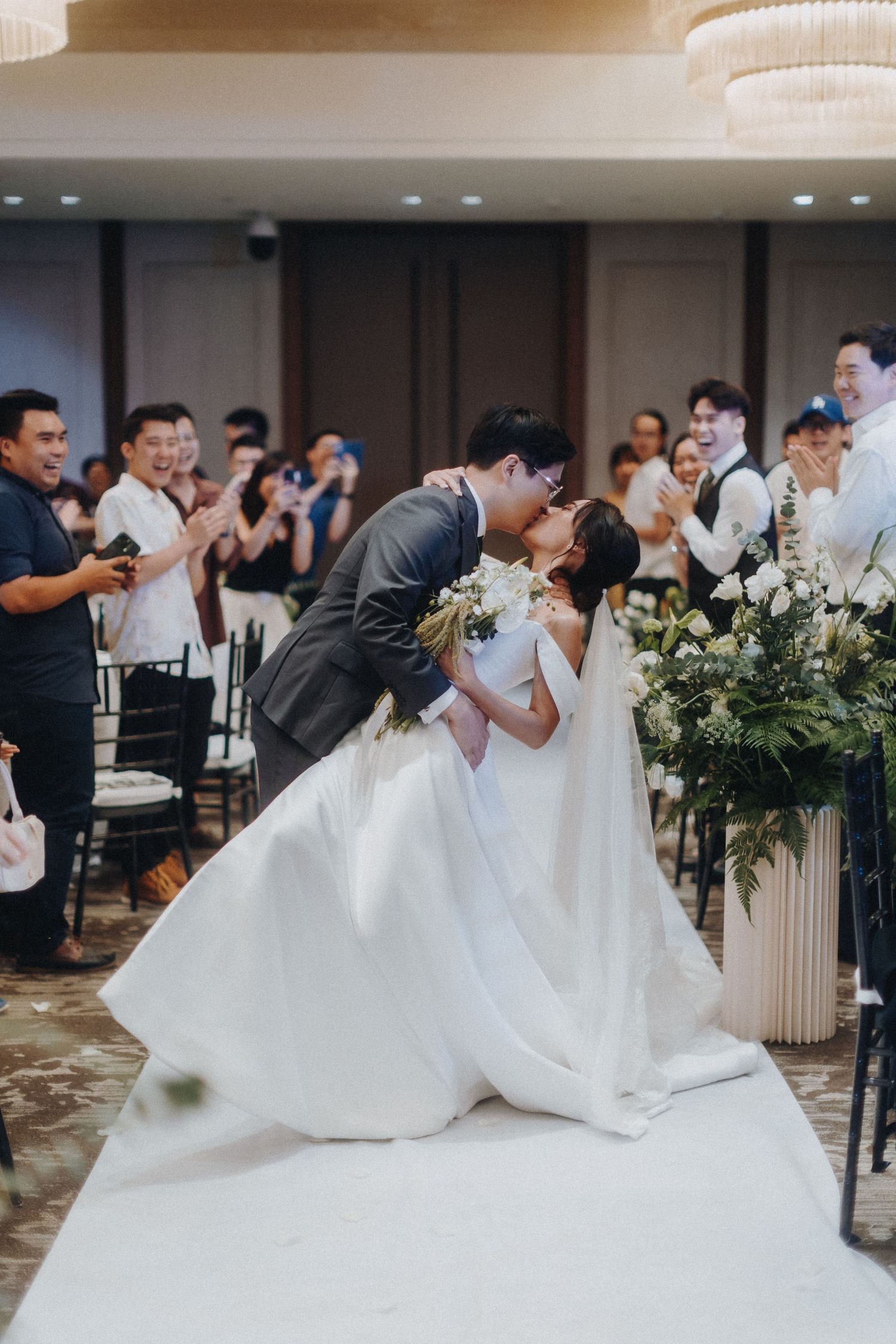 Newlyweds share their first kiss during indoor wedding ceremony with floral decorations.
