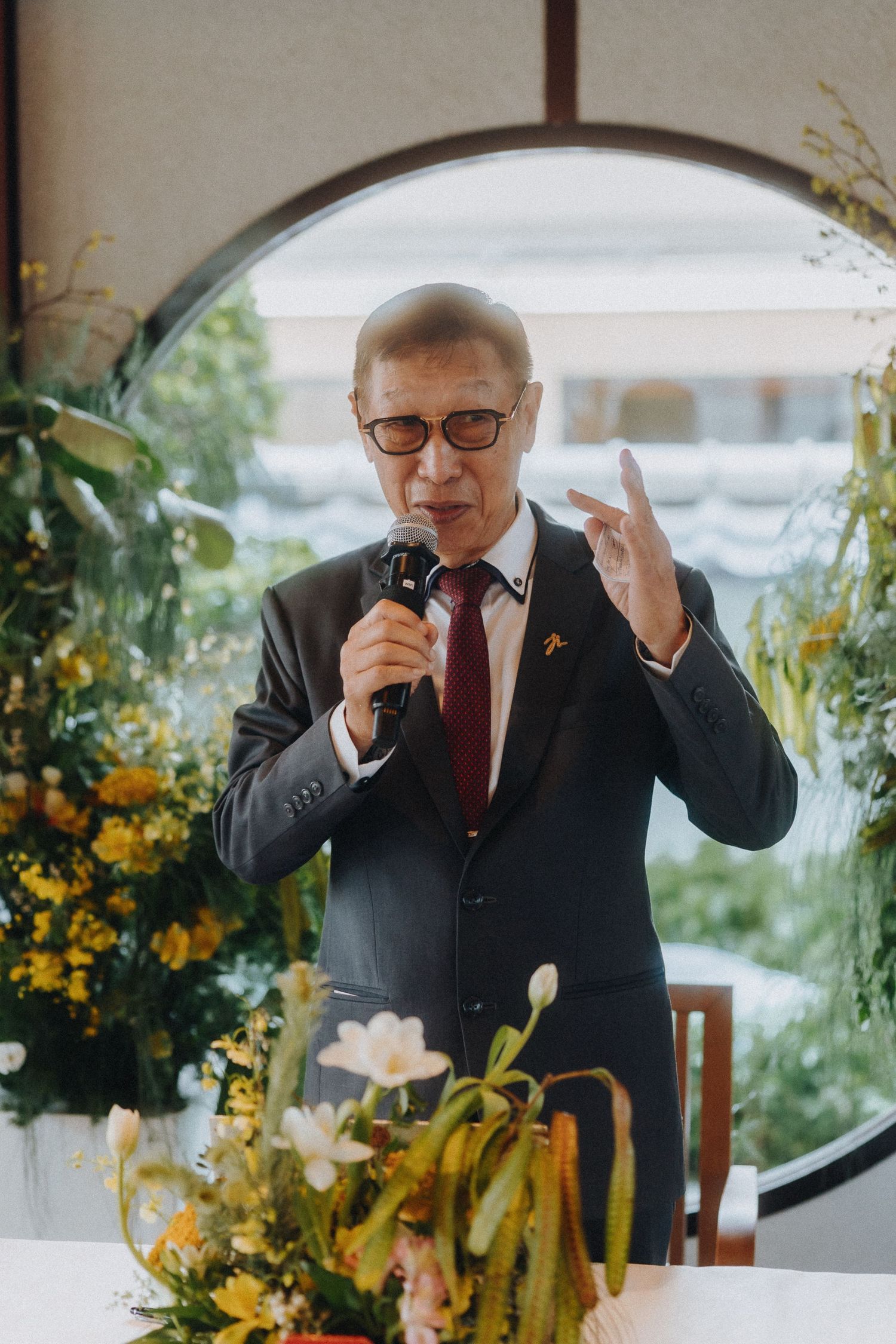 A speaker in a dark suit and glasses gives a speech at an event surrounded by floral decorations.