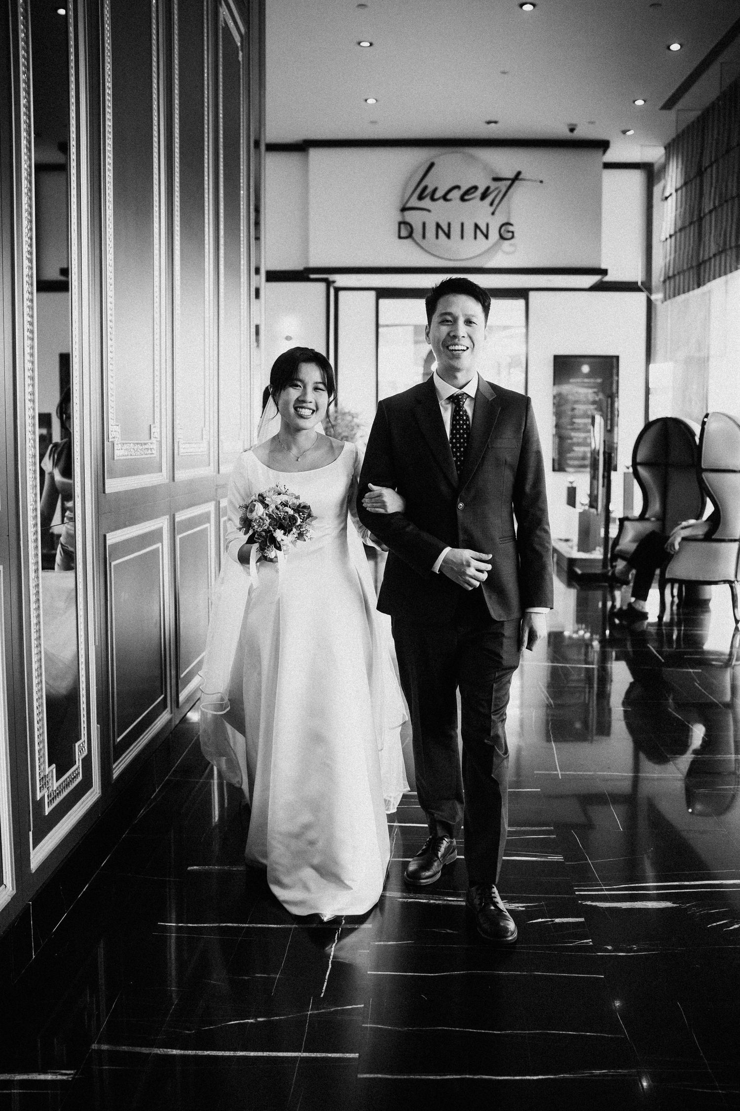 Happy bride and groom walk arm in arm through Lucent Dining venue in classic black and white wedding photo.
