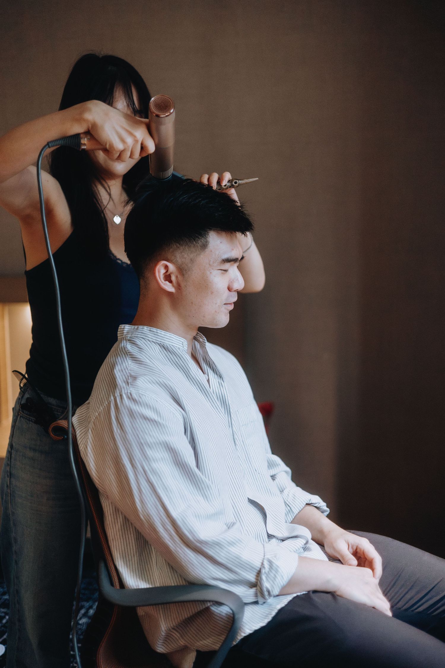 A hairstylist uses scissors and a hair dryer to style a young mans short dark hair during a wedding preparation session.