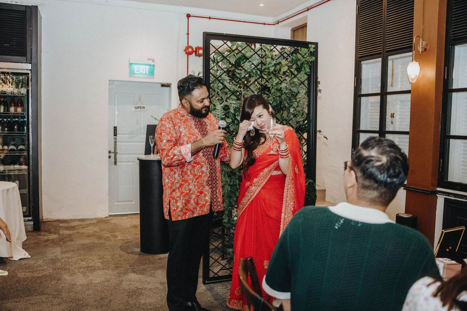 Couple in traditional Indian wedding attire interact with guests inside elegant venue.