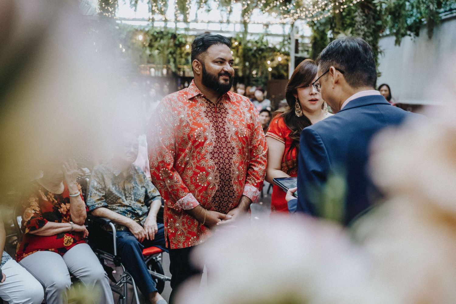 Man in red floral batik shirt greets guests at an elegant outdoor multicultural wedding event.