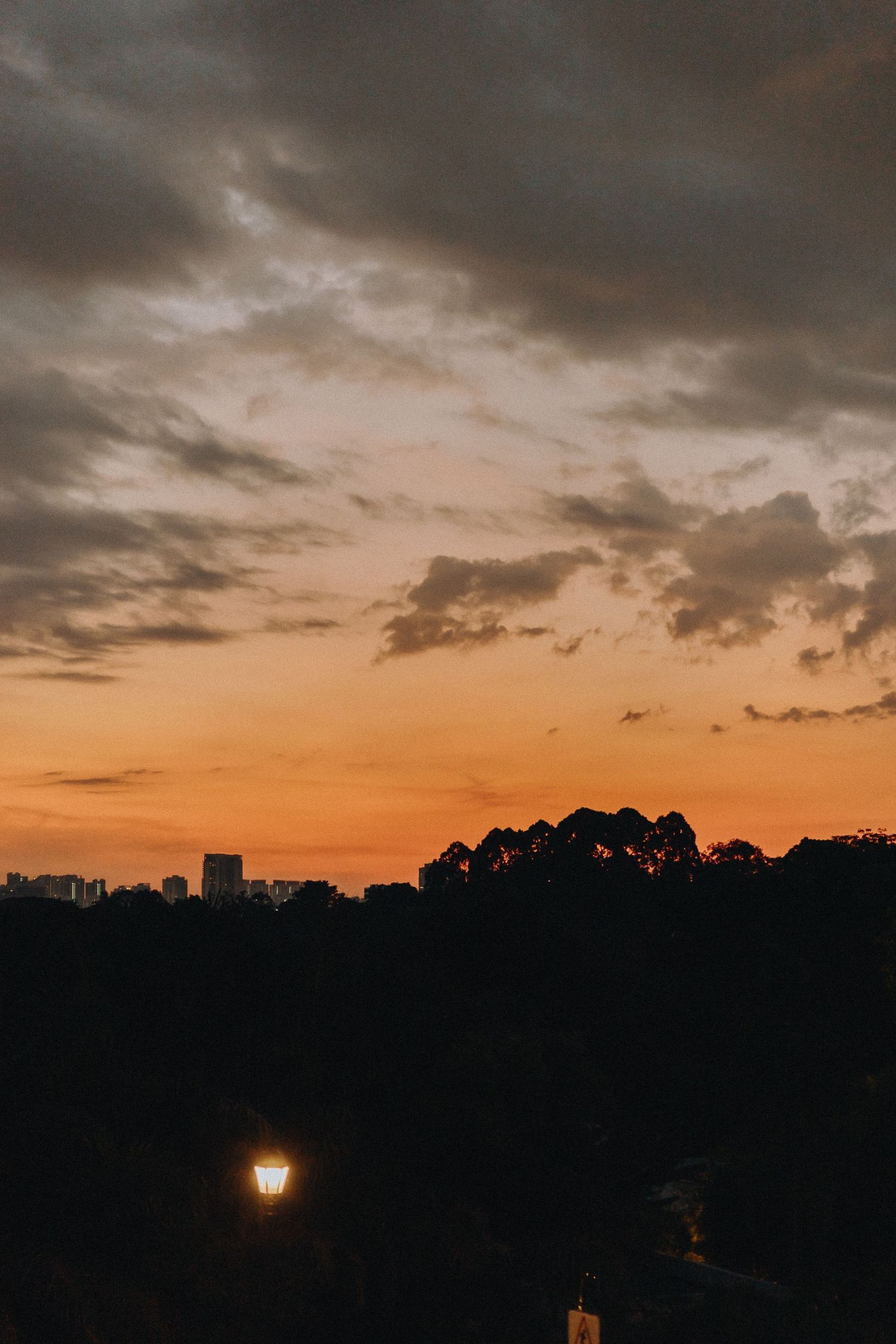 Dramatic sunset sky over city skyline with single illuminated street lamp in dark foreground.
