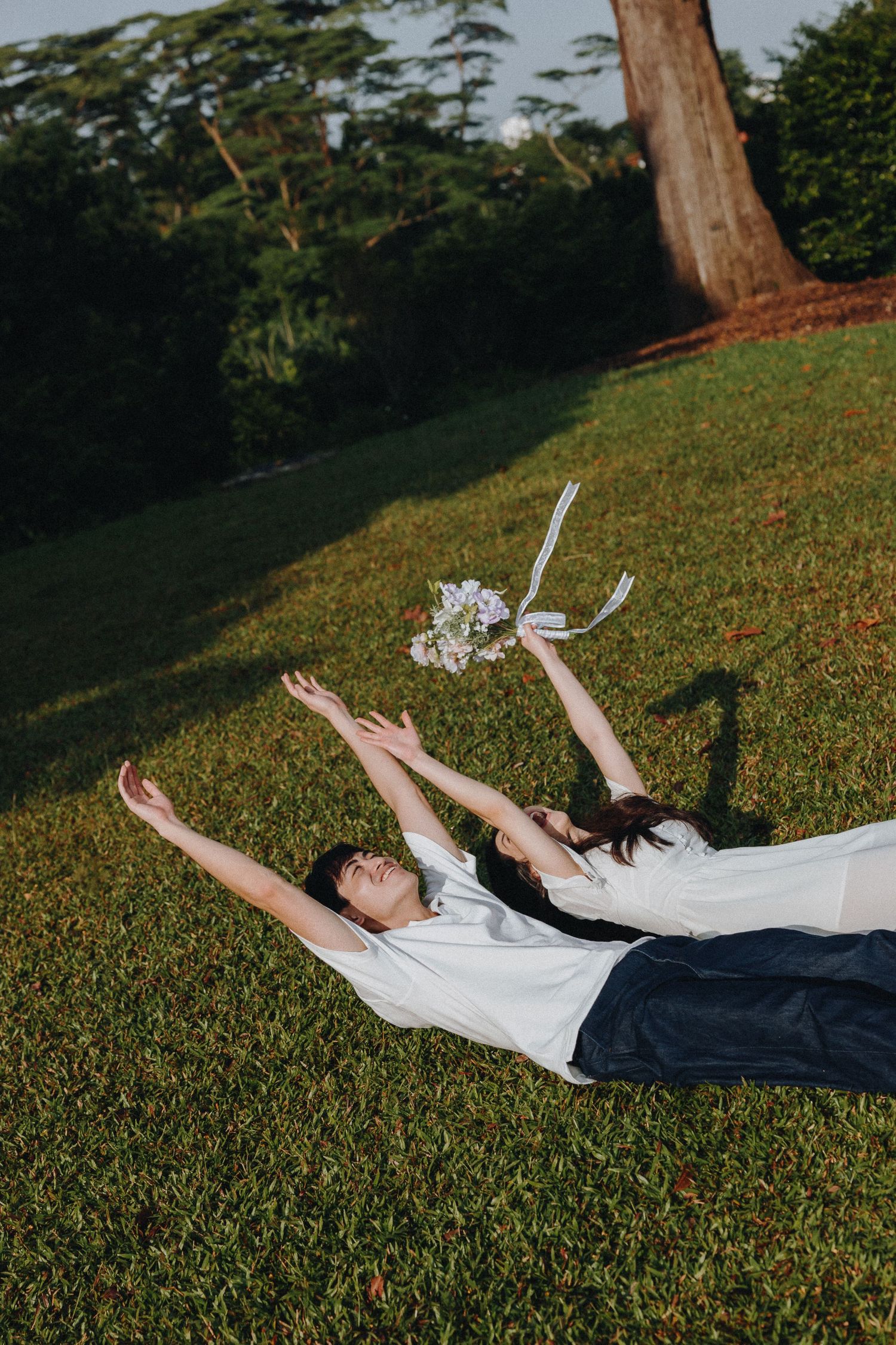 Two people lying on grass holding bouquet up in playful pose.