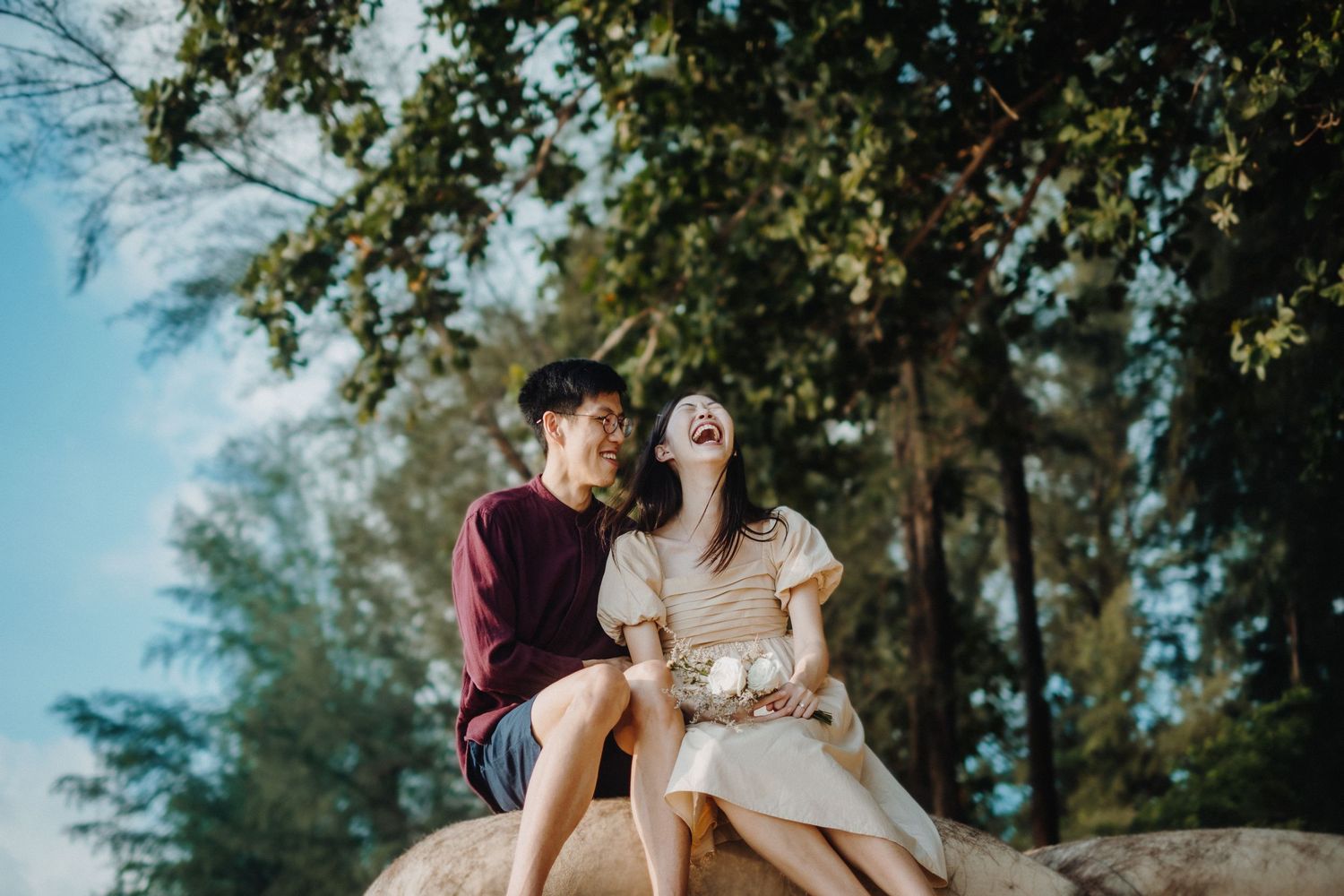 A couple sits together on a stone ledge under green tree branches on a sunny afternoon.