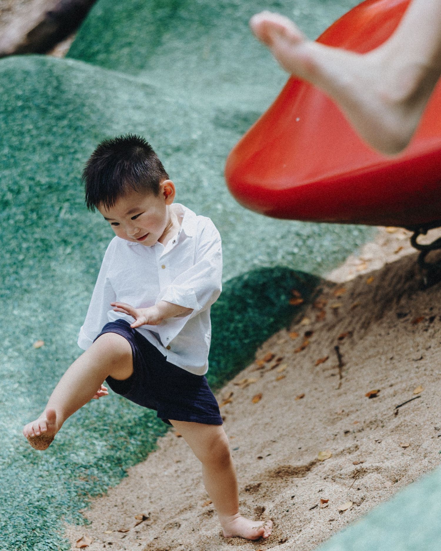 A young child in a white shirt and navy shorts plays barefoot on a playground with a red slide visible.