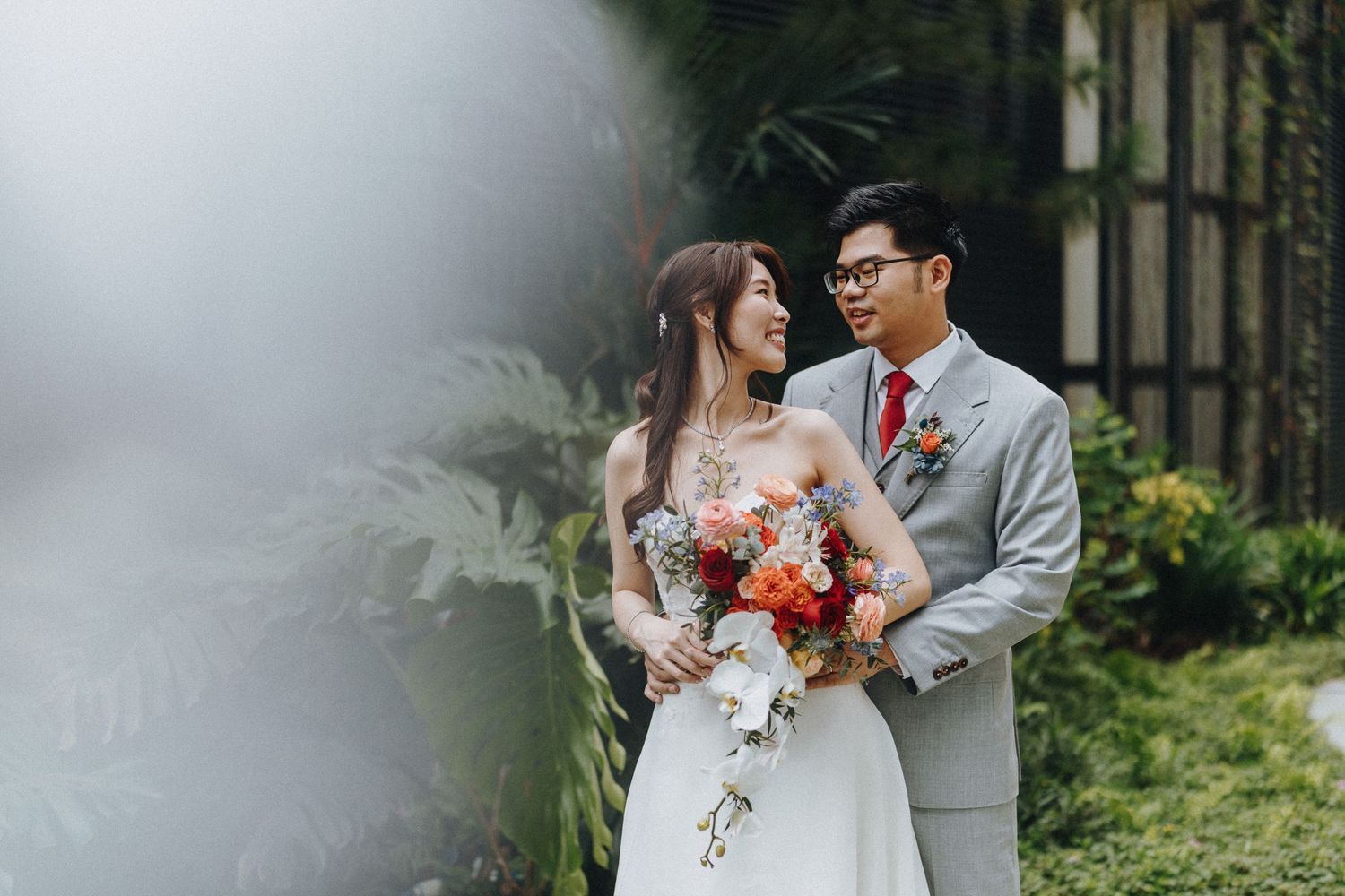 A couple stands together in a garden setting, the bride holding a vibrant bouquet while wearing an elegant white gown.