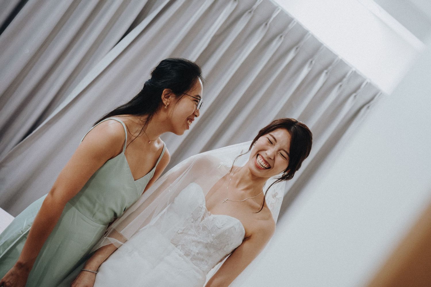 Two people share a joyful moment during wedding preparations in a bright interior room with white curtains.