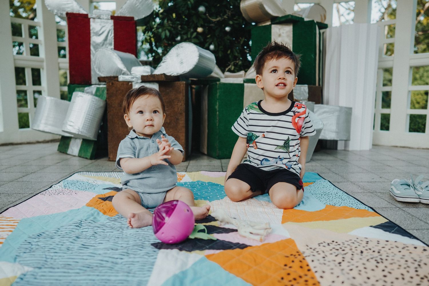 Two young children sit together on a colorful quilt while playing and laughing on a porch.