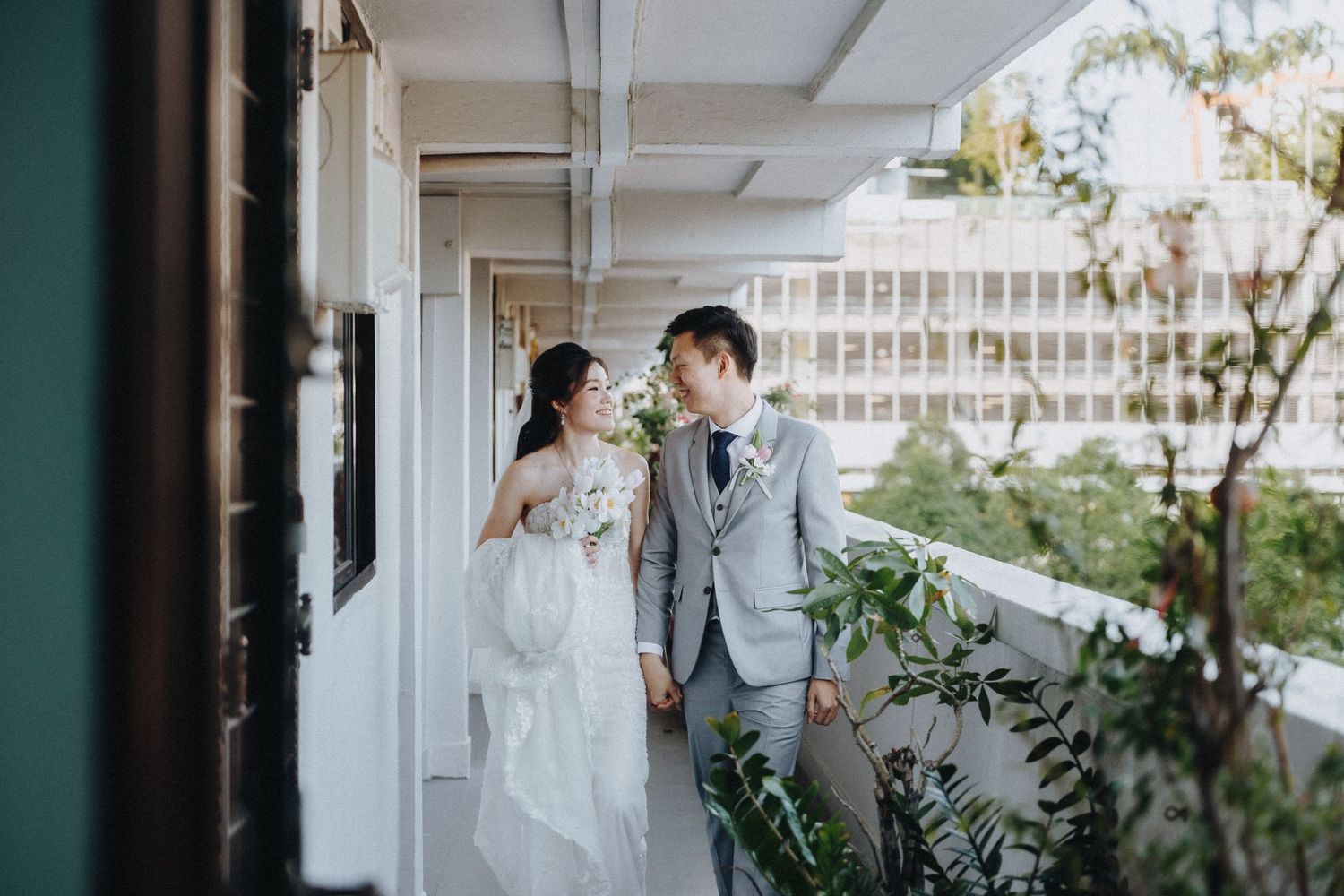 A couple in wedding attire smiles at each other while walking along an outdoor corridor with greenery.