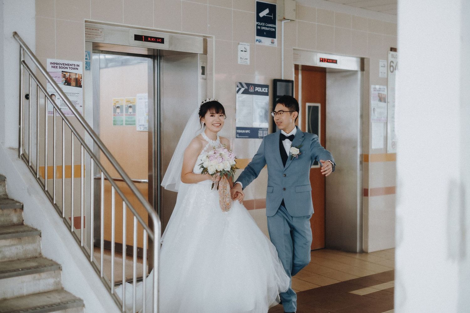 A wedding couple walking down stairs near elevators in a bright indoor setting.