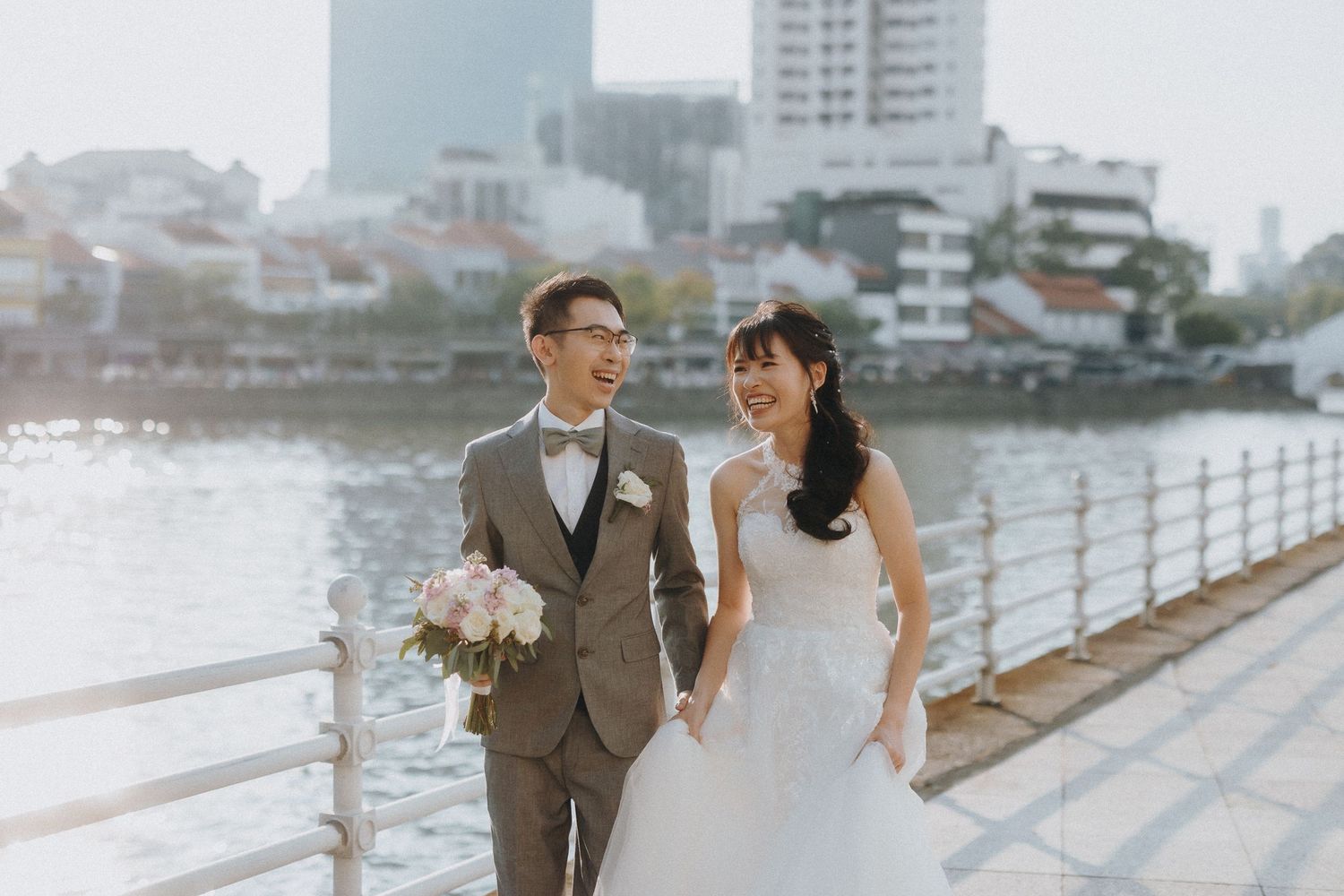 Couple in wedding attire taking photos along urban waterfront promenade.