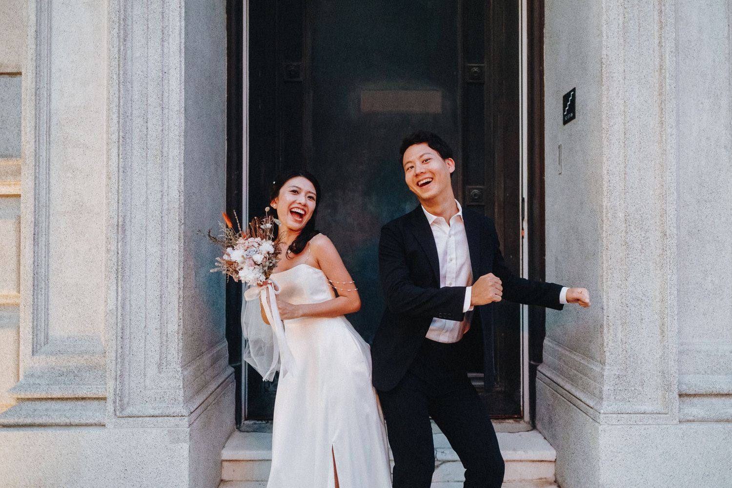 Wedding couple posing playfully between white classical columns in formal attire.
