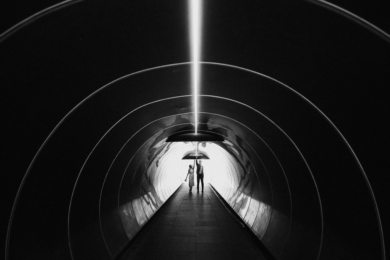 Black and white photo of figures with umbrella walking through illuminated circular tunnel.