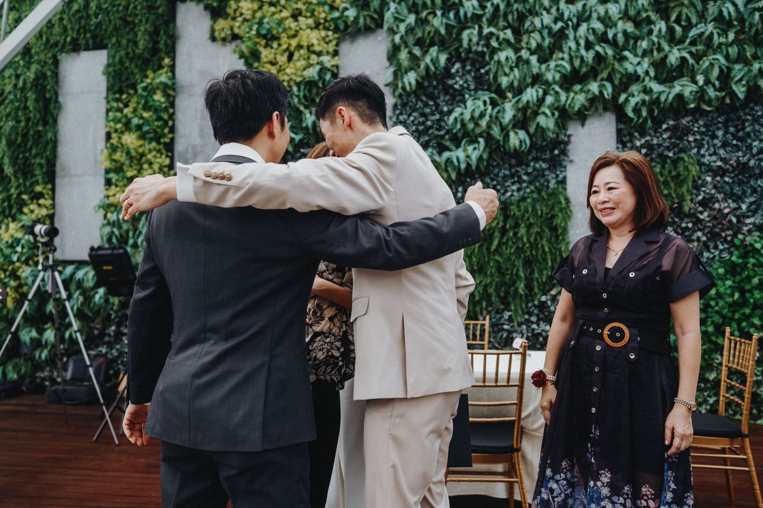 Wedding guests embrace during an emotional moment at an outdoor reception with ivy-covered walls.