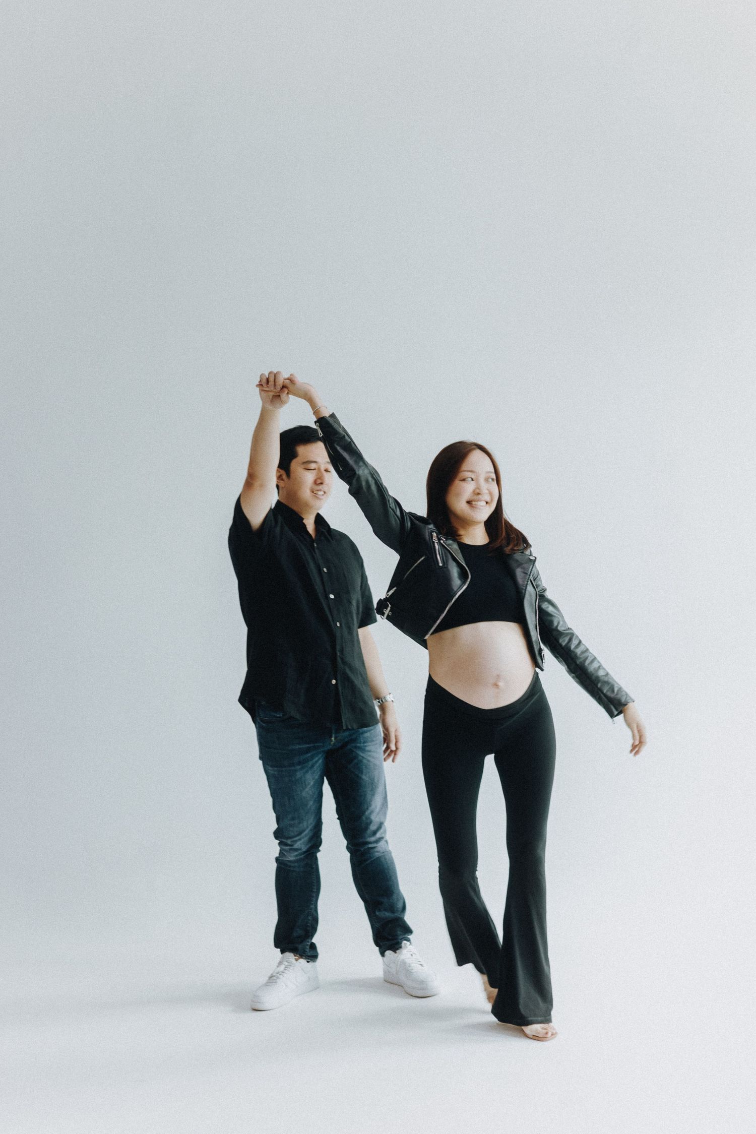Expectant couple dances together against white backdrop while holding hands up in celebration.