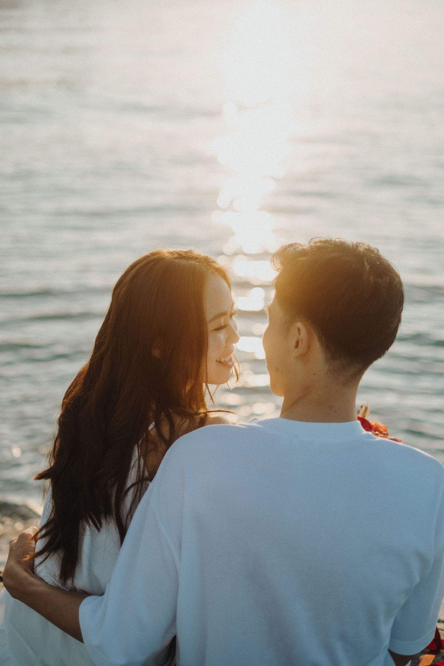 A couple shares an intimate moment by the ocean with golden sunlight reflecting on the water.
