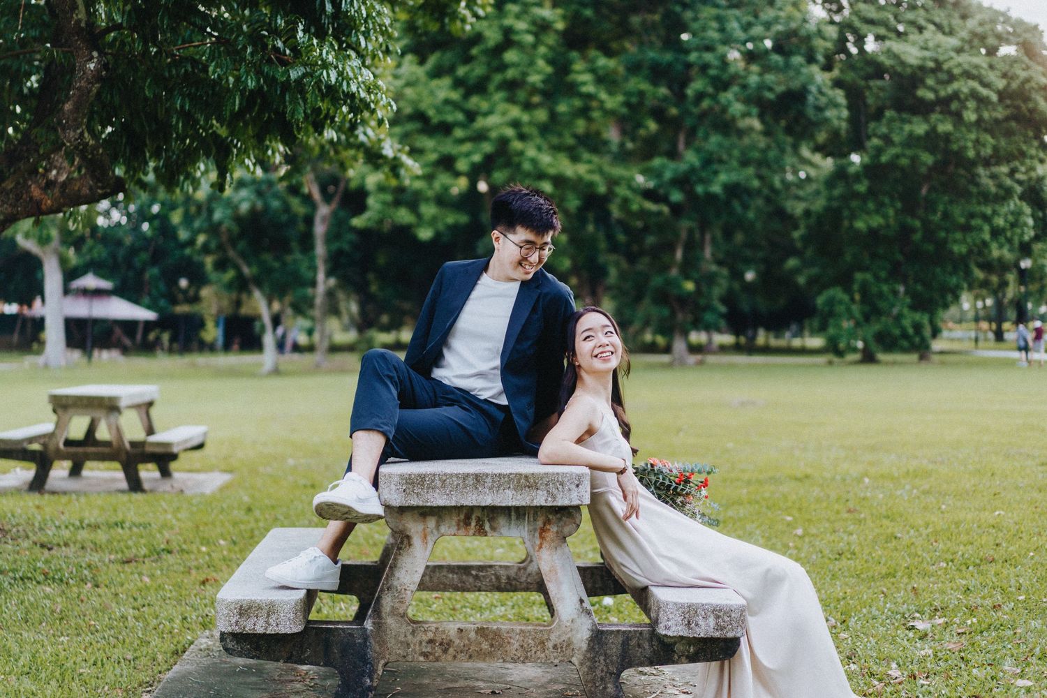 A couple in wedding attire sits together on a wooden picnic table in a serene park setting surrounded by greenery.