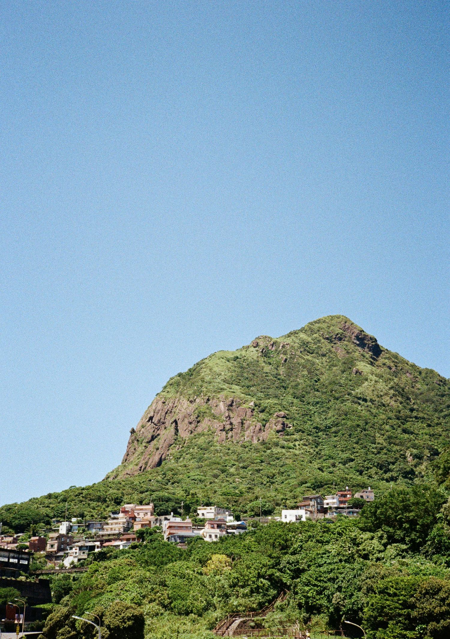 Houses perched along a green mountainside with rocky cliffs and blue sky in a scenic coastal landscape.