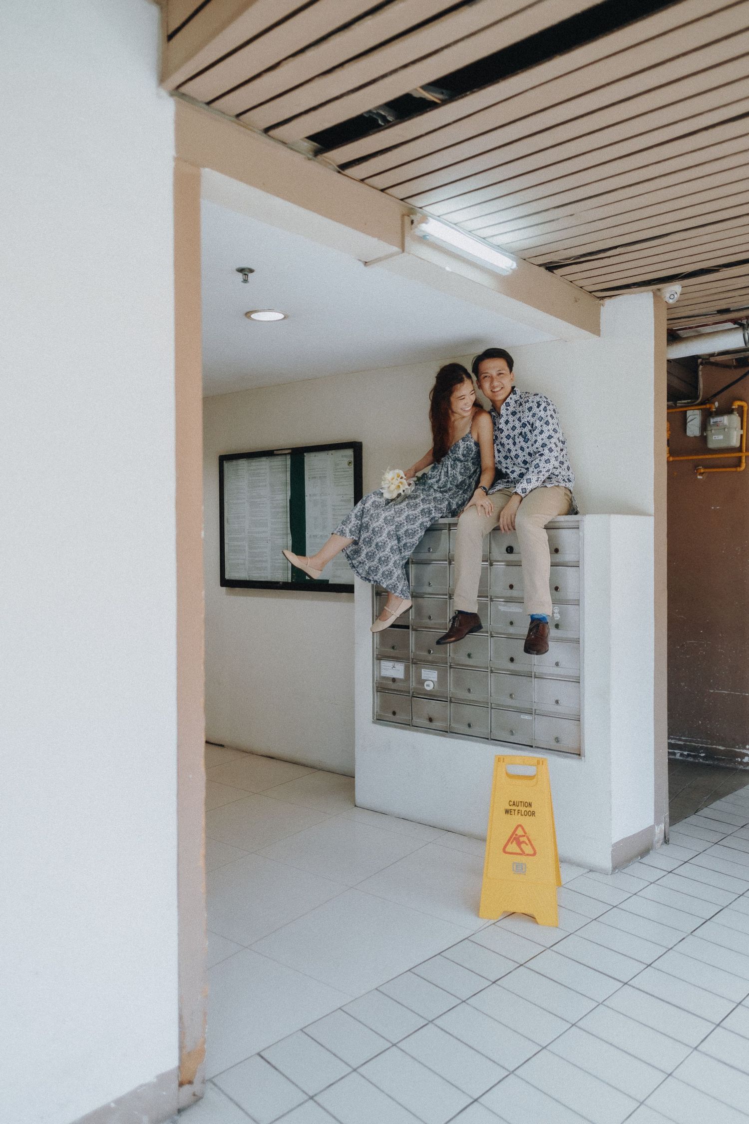 A couple sits playfully on concrete steps in a modern indoor space with white walls and tiled floors.