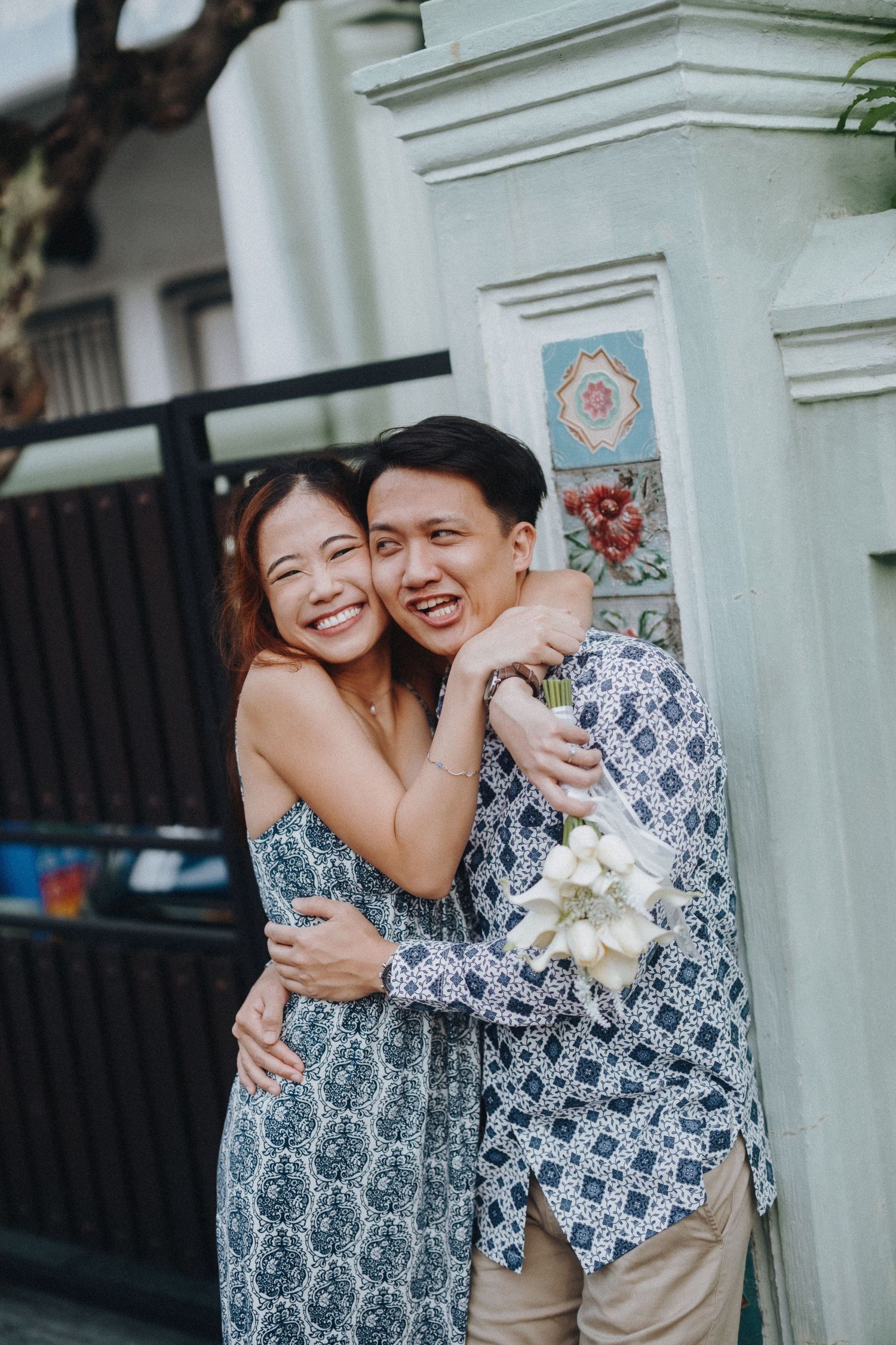 A couple in matching blue and white patterned outfits share joyful embraces and laughter against a white wall.