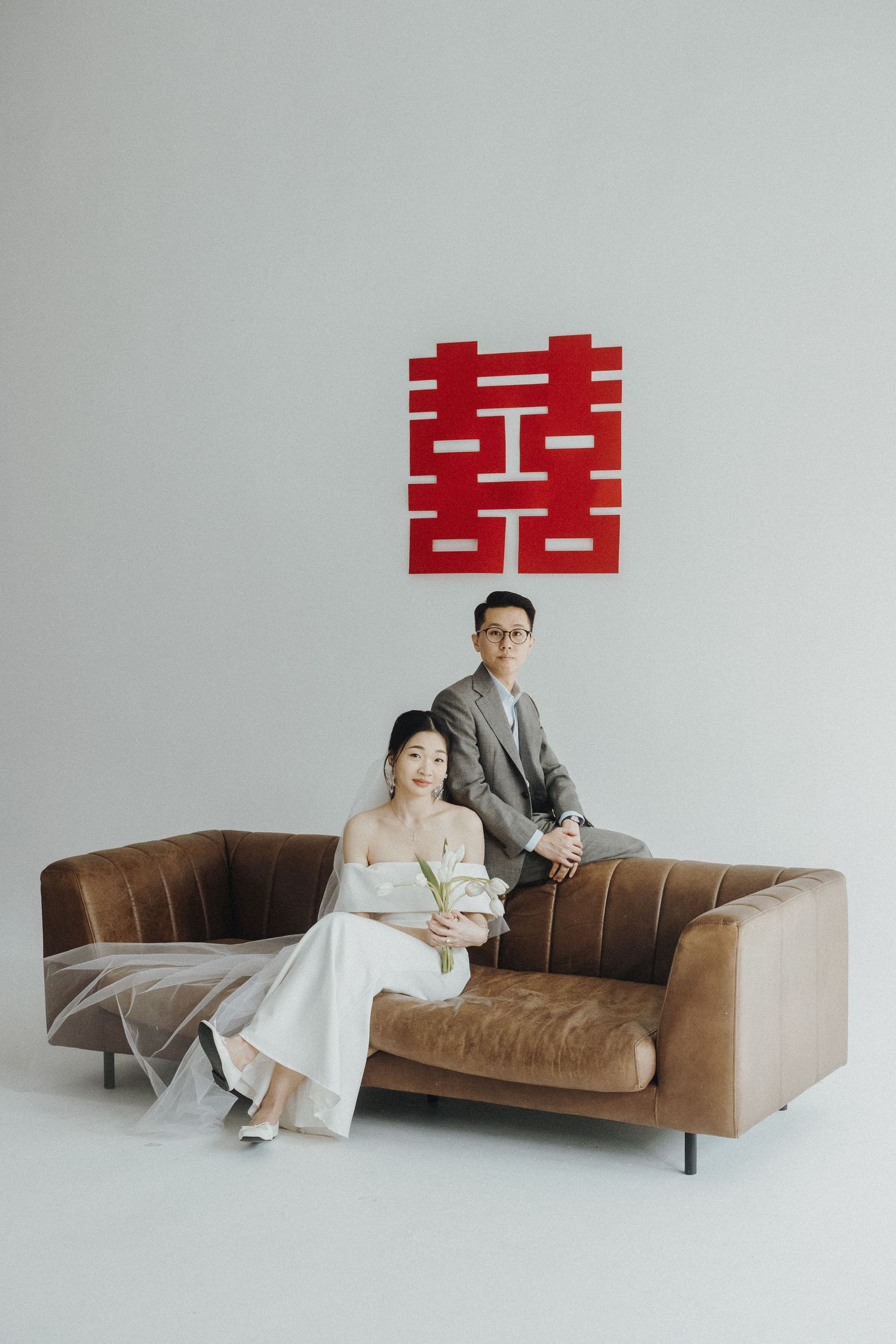 Wedding couple poses on a brown leather couch in a minimalist studio setting with white walls.