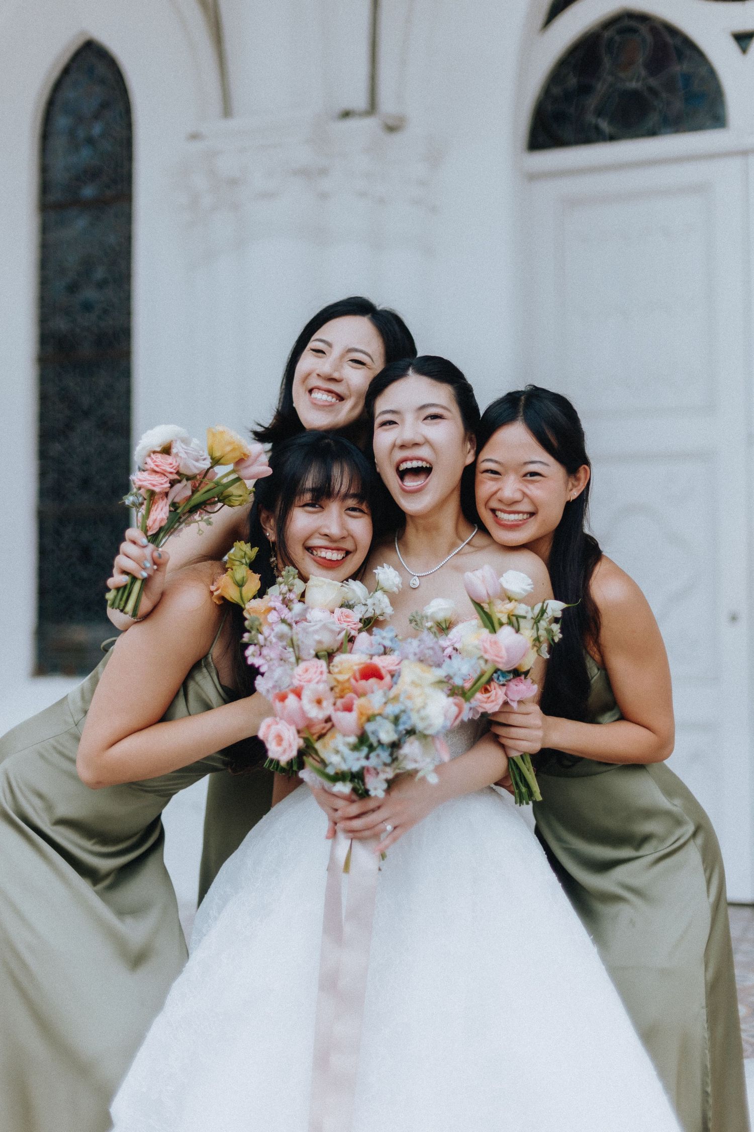 Bride in white gown laughing joyfully with three bridesmaids in olive green dresses holding colorful bouquets.