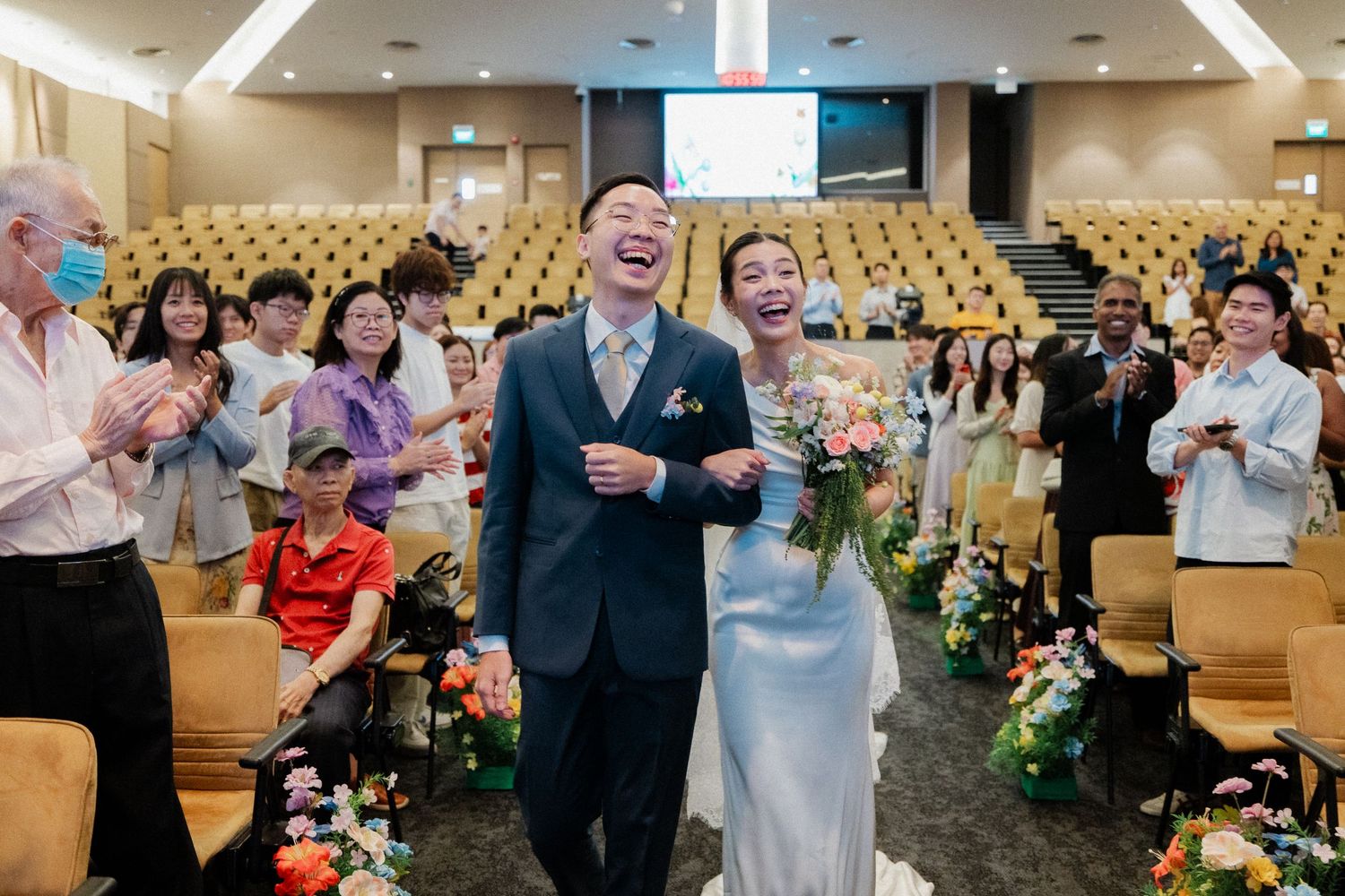 Joyful newlyweds walk down aisle laughing at wedding ceremony with guests applauding.