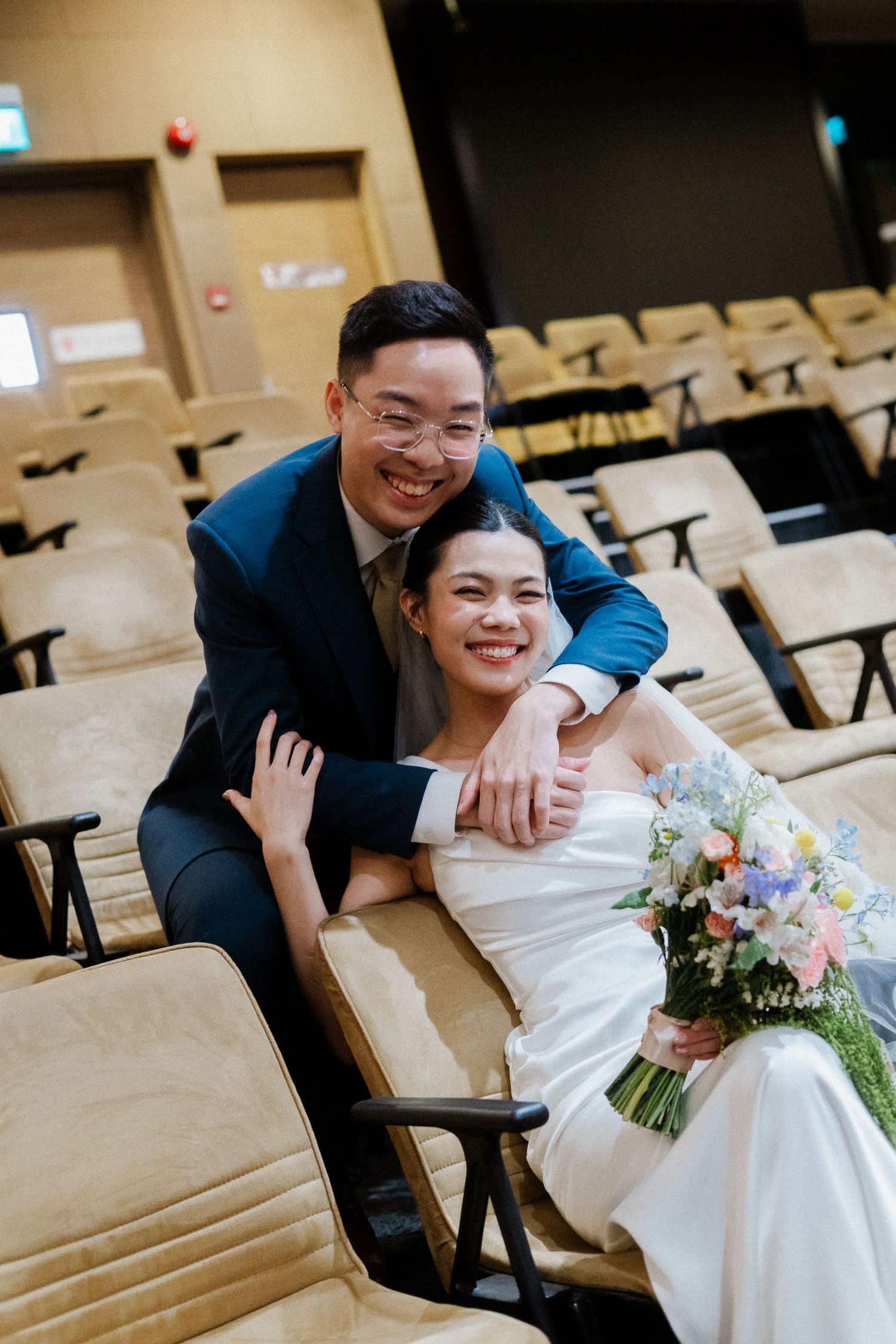 Smiling bride and groom pose together in auditorium seats, bride holding colorful bouquet in white gown, groom in blue suit.