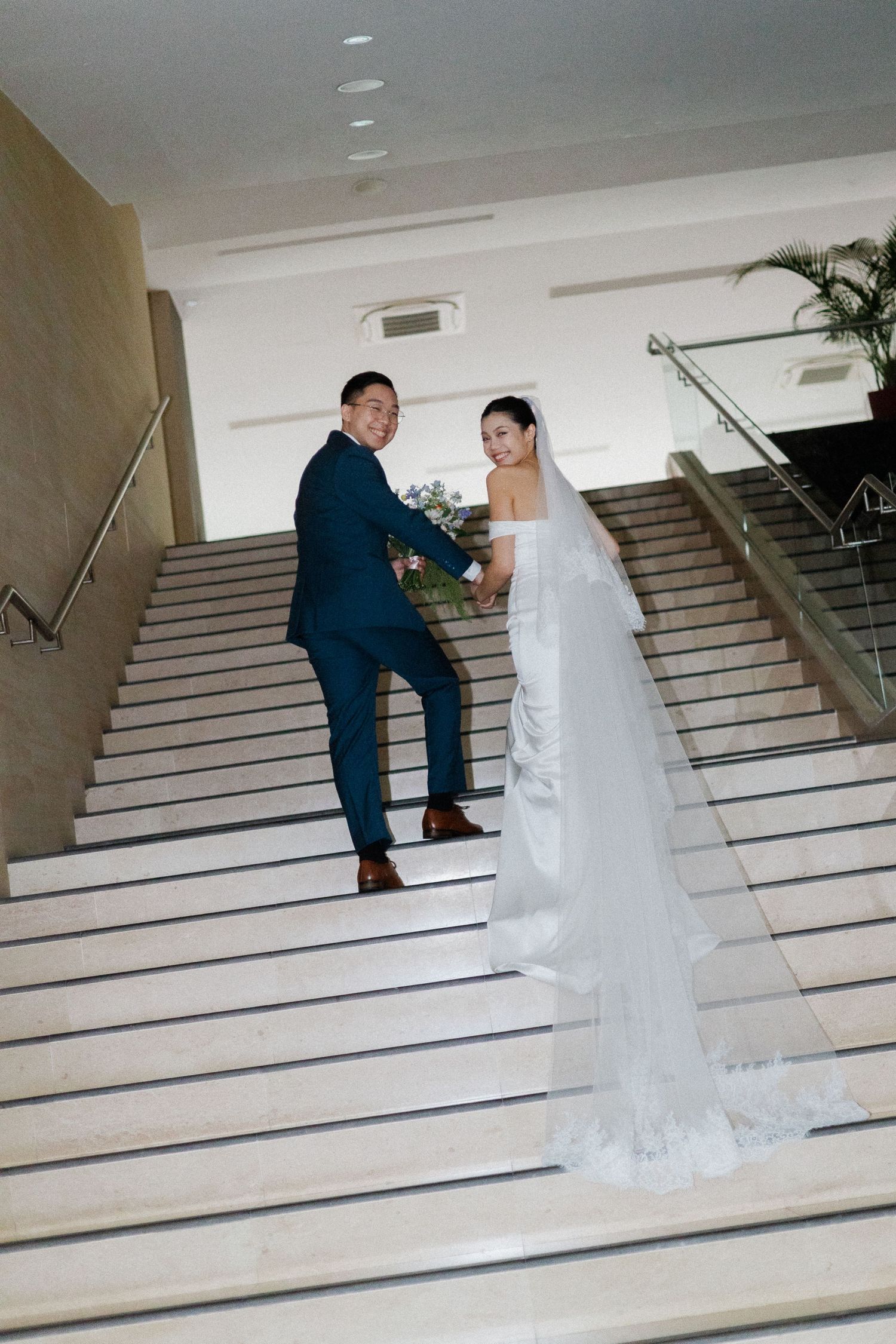 Bride and groom posing on grand staircase, bride in white gown with veil, groom in teal suit holding bouquet.