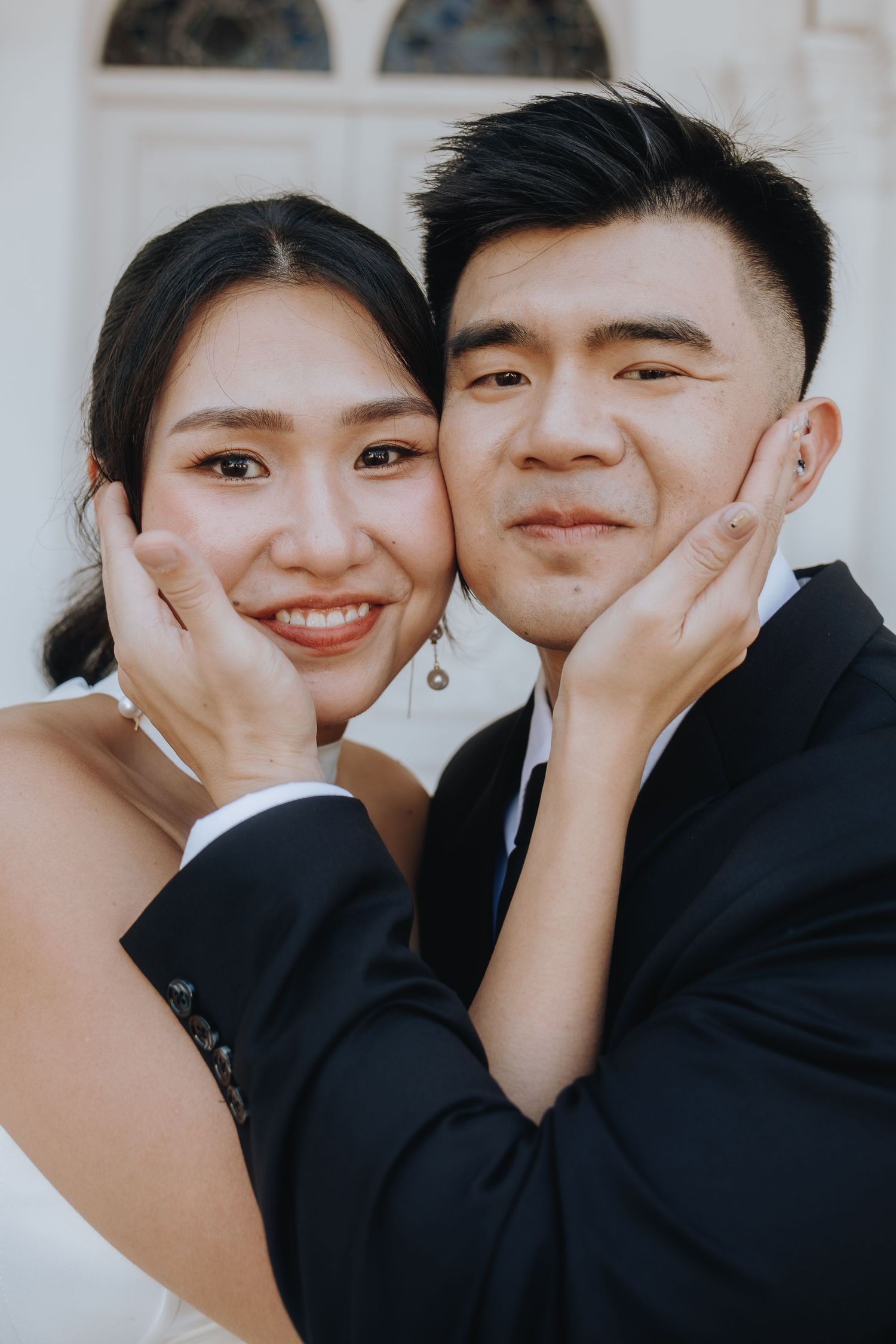 Smiling Asian bride and groom posing closely together outside a white building.