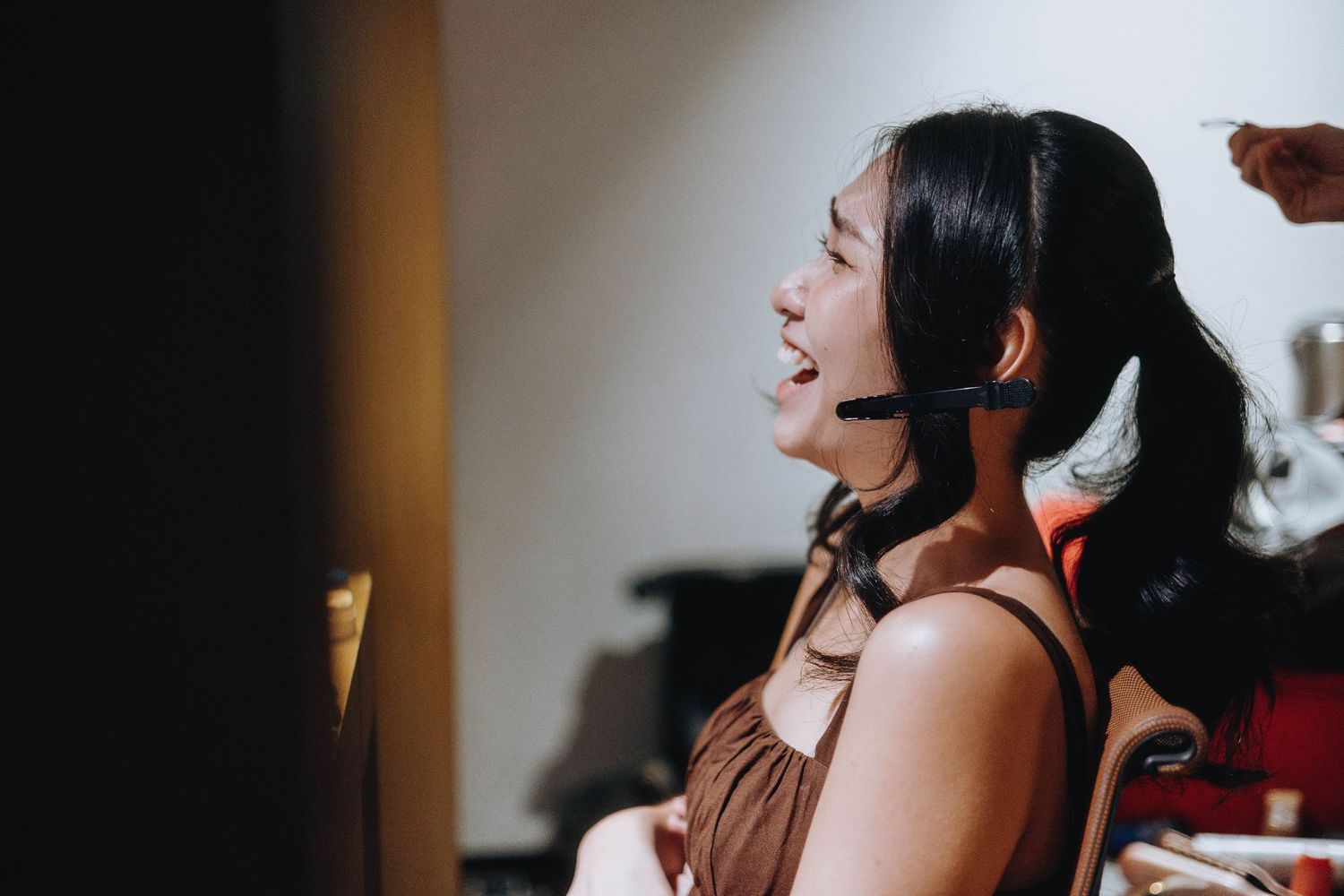 Young Asian woman with headset mic singing passionately, wearing brown dress, side profile view indoors.