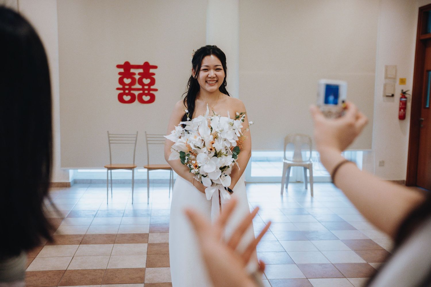 Bride in white gown holds floral bouquet while being photographed by guests at her wedding reception.