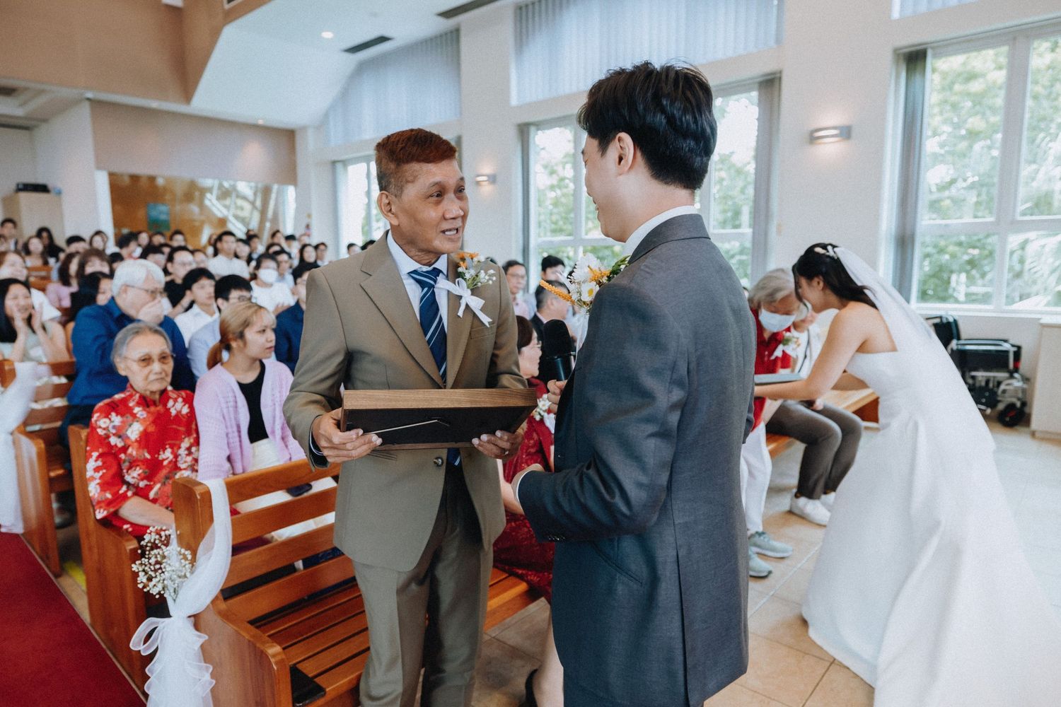 Groom receives a gift from a guest during wedding ceremony while bride waits in the background.