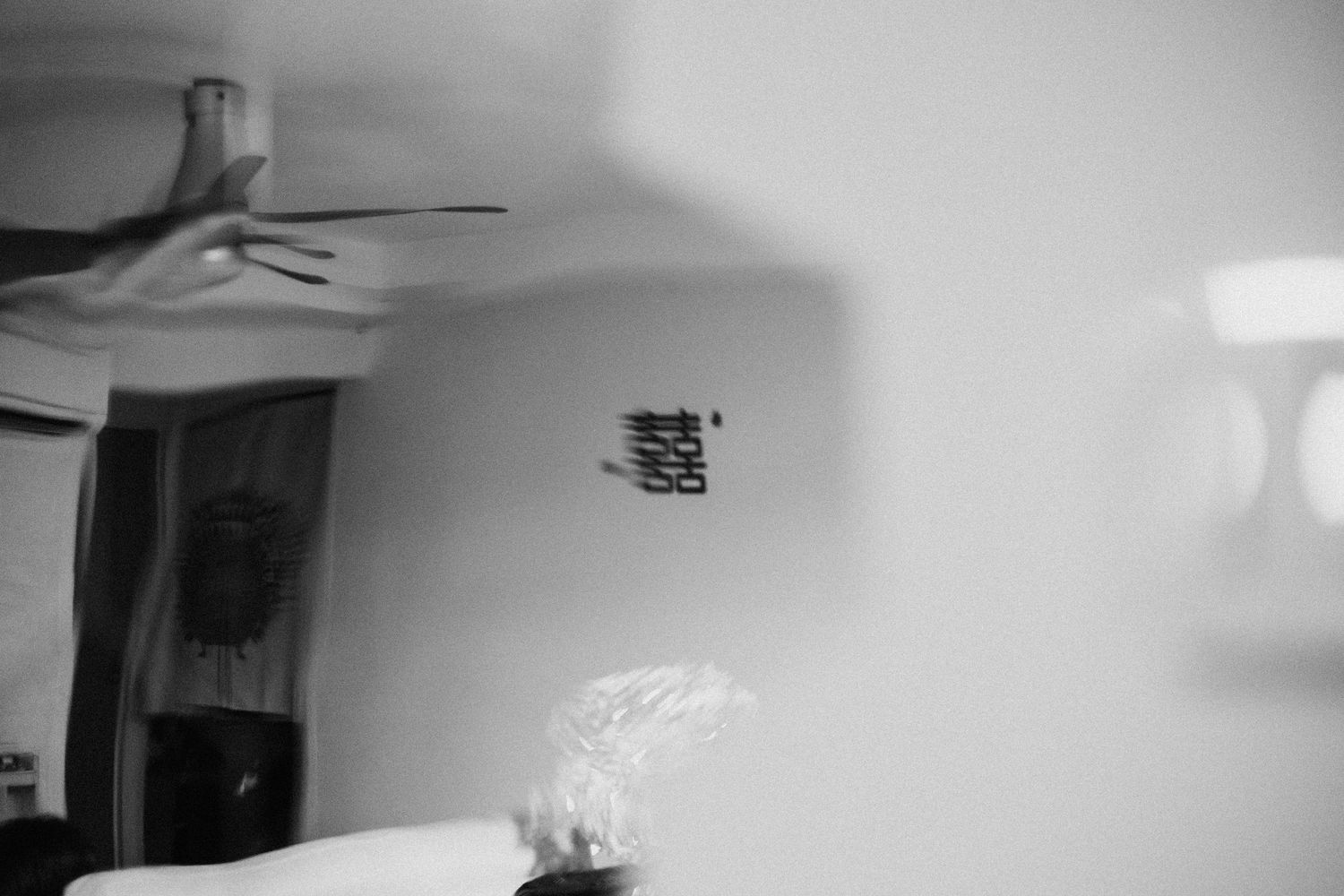 Black and white photo of a bedroom with ceiling fan, wall decor, and soft lighting on the right side.