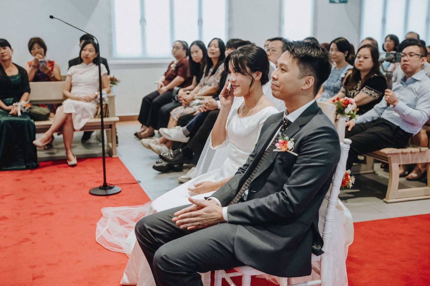 Asian bride and groom seated at wedding ceremony with guests in background.
