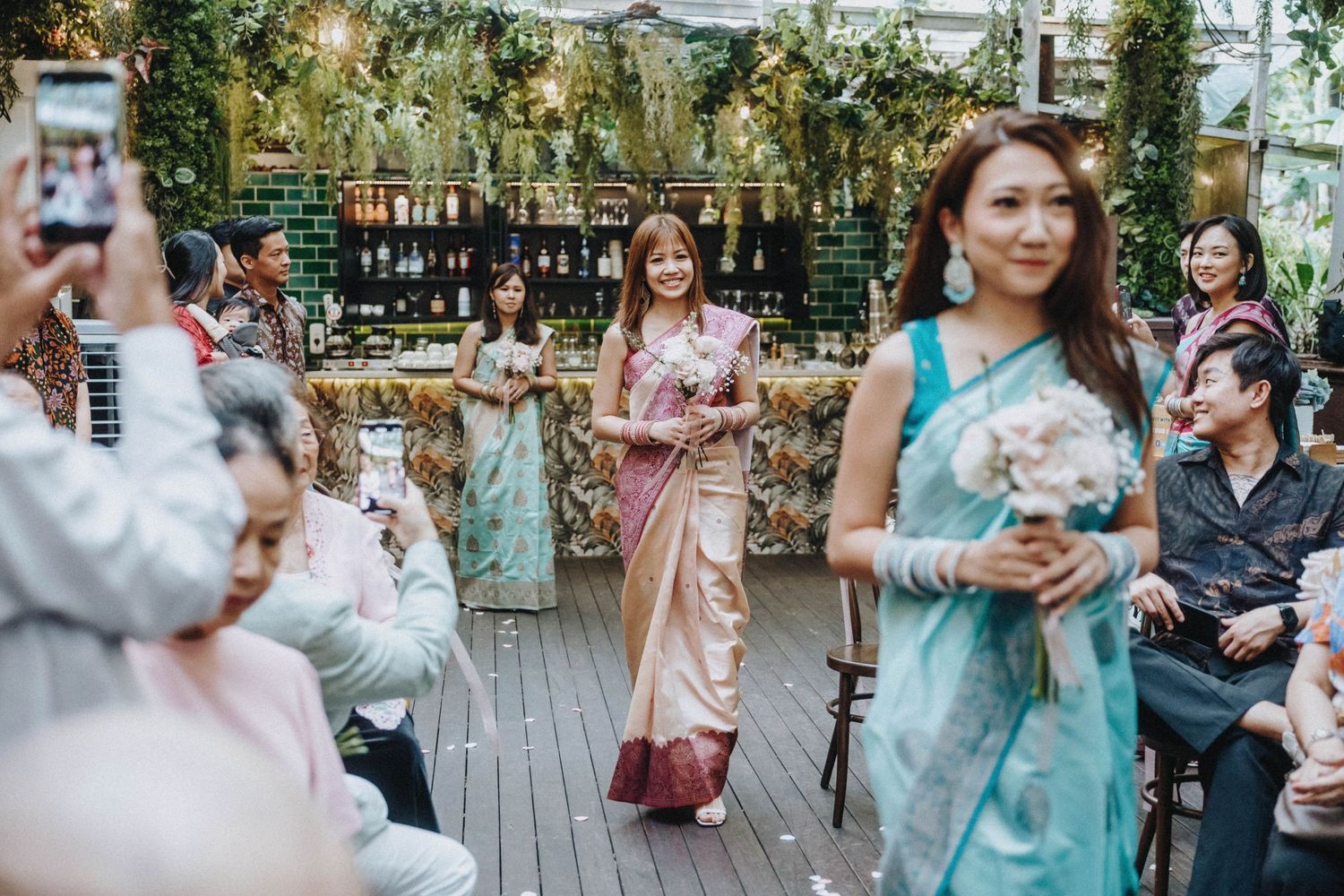 Bridesmaids in traditional Thai silk dresses walking down the aisle at an outdoor garden wedding ceremony.