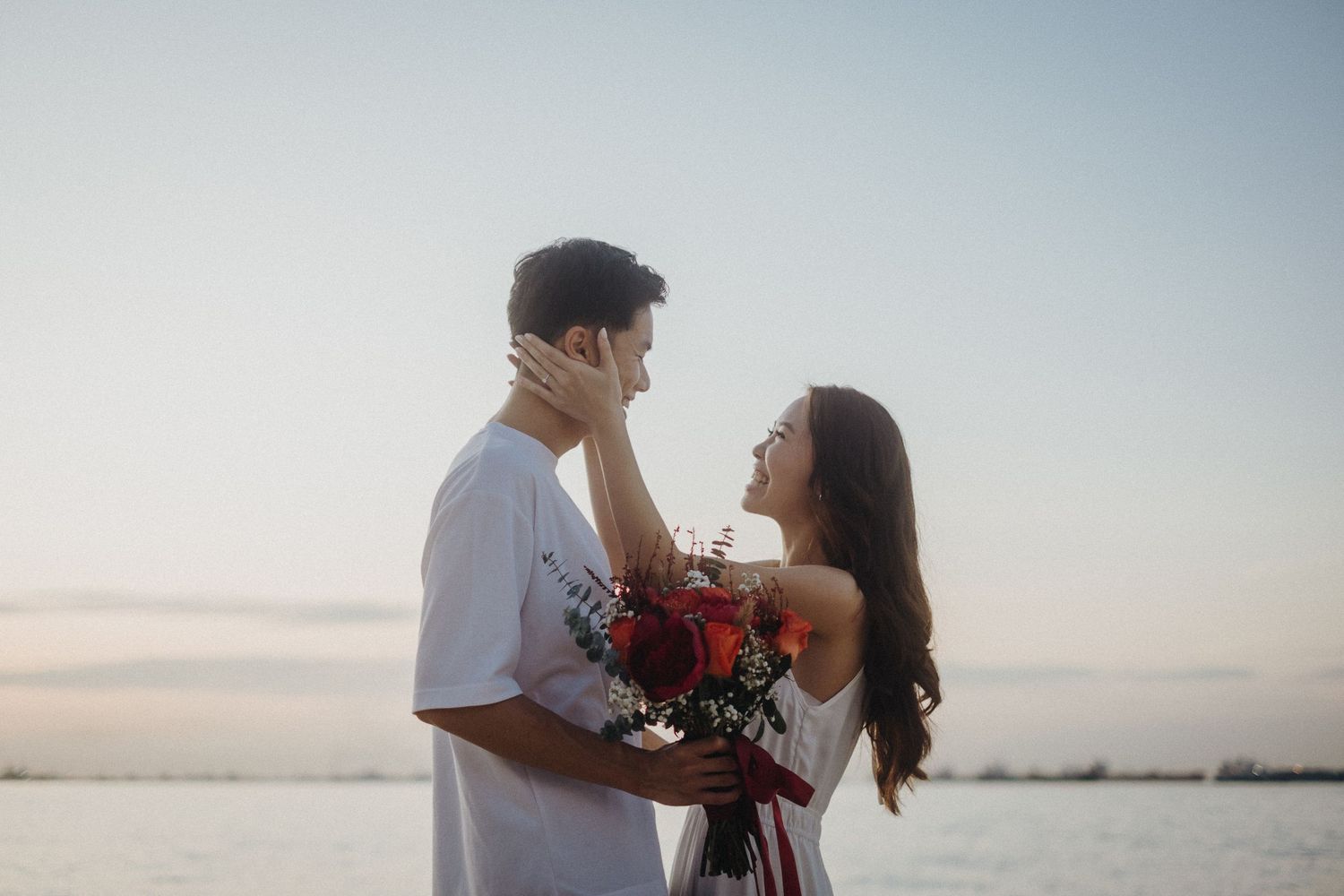 Two people embracing on a beach during sunset with ocean waves and distant mountains in the background.