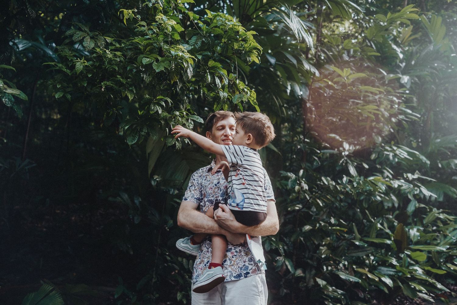 Parent lifts child in playful moment against lush green foliage background.