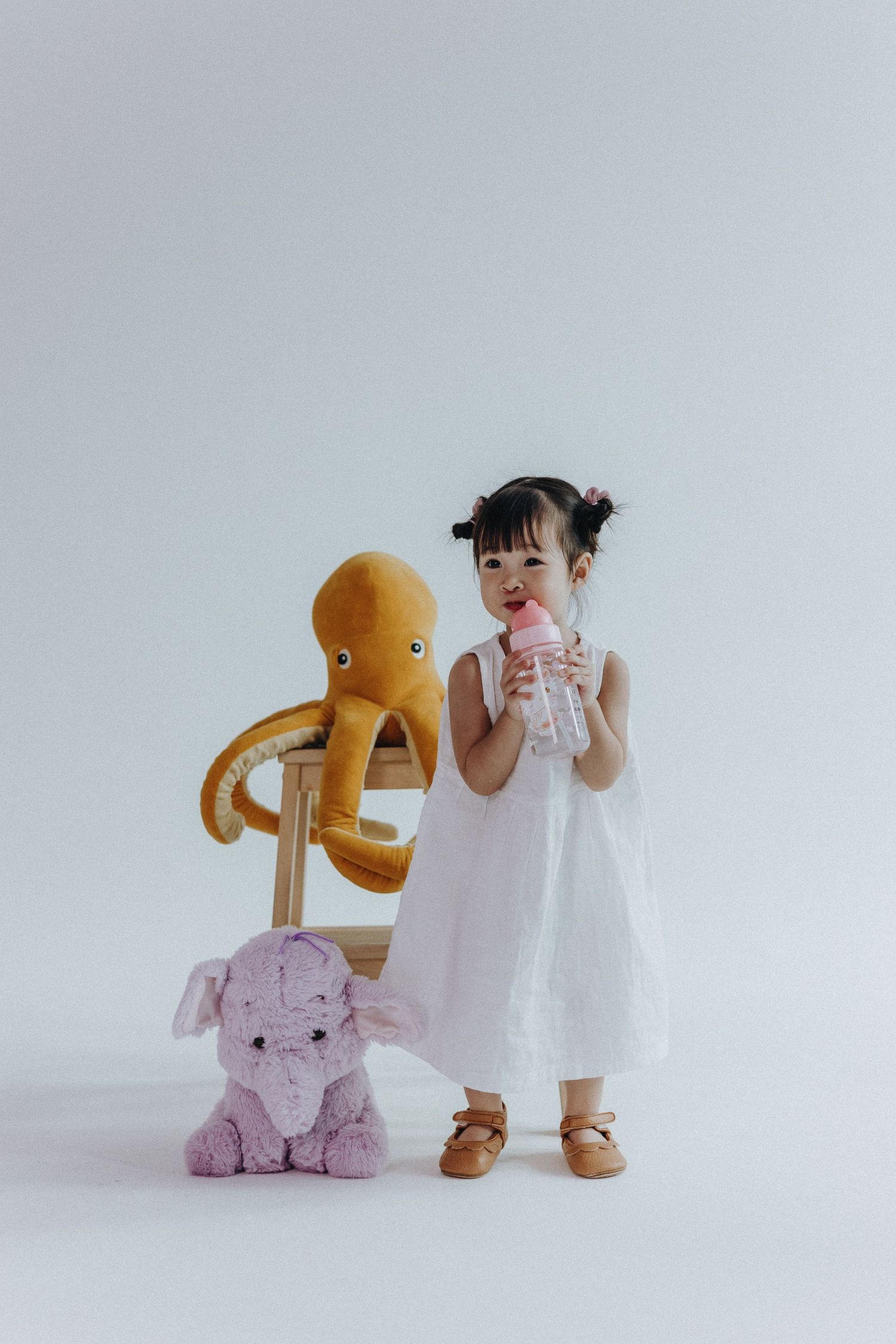 Child in white dress plays with plush toys including orange and pink stuffed animals.