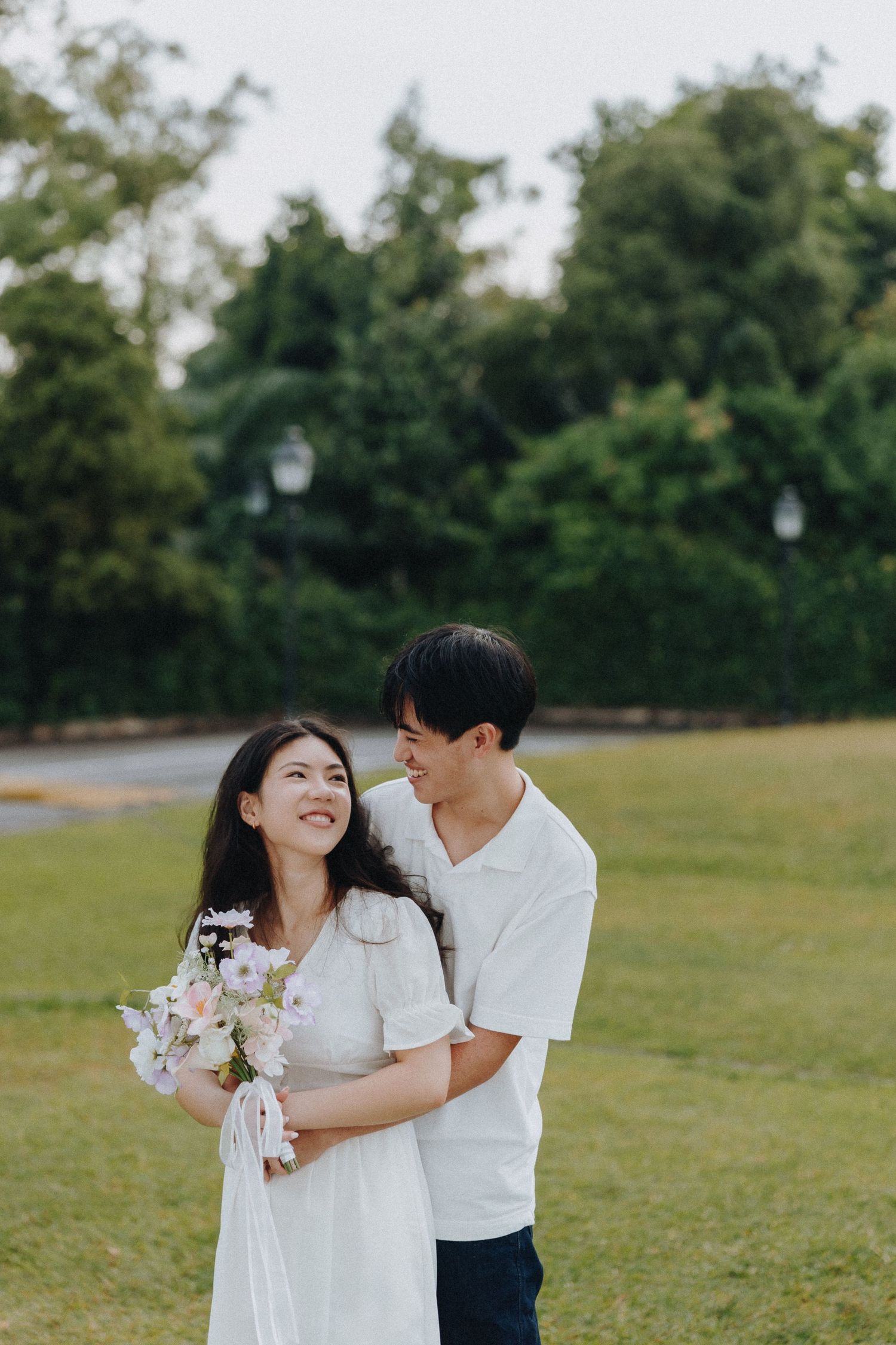 A couple in white outfits sharing a tender moment in a green garden while holding a flower bouquet.