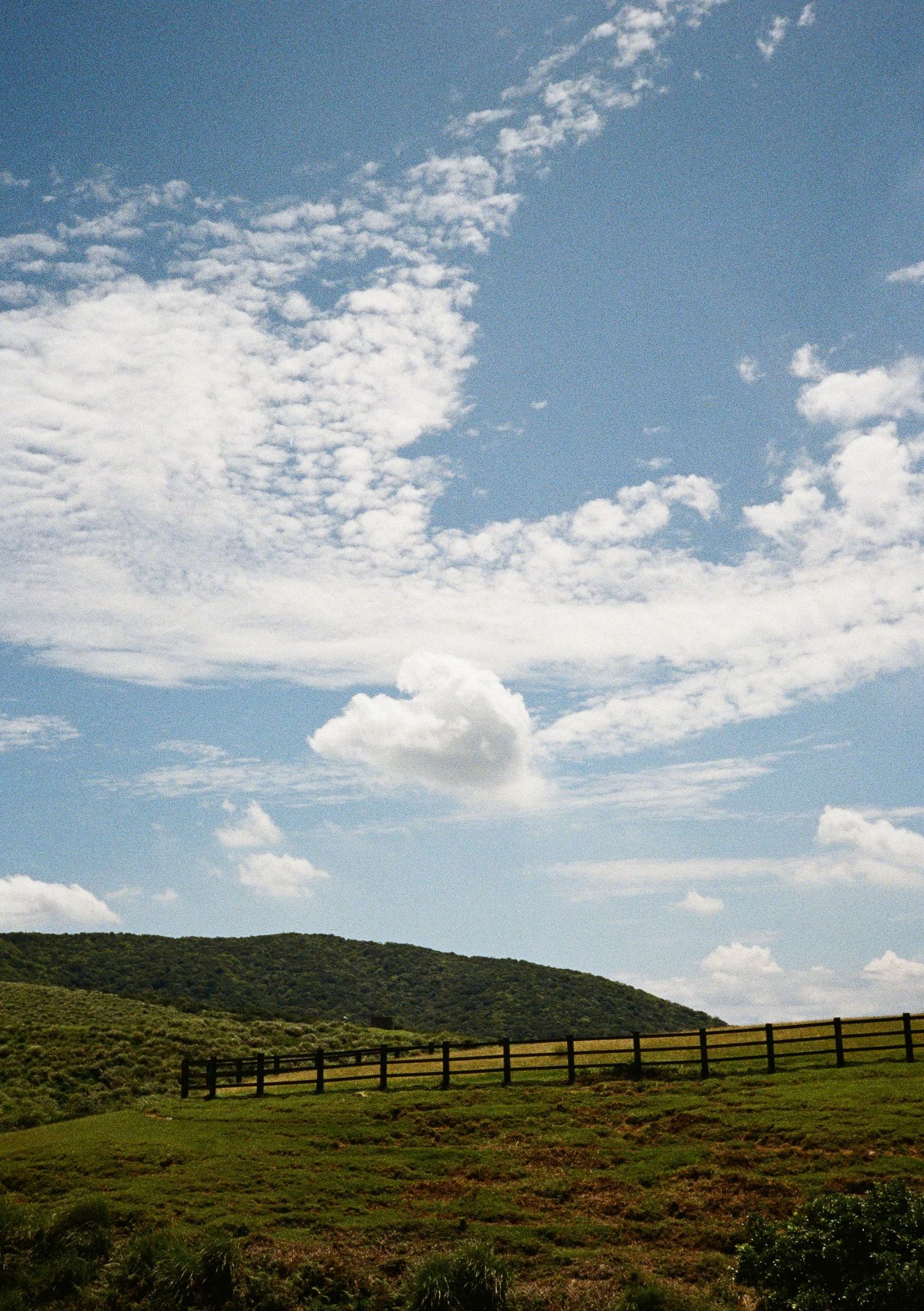 Scenic hillside with wooden fence running along the ridge under scattered clouds.