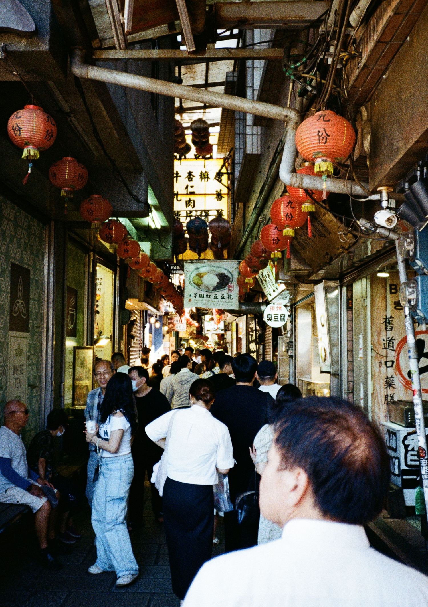 Traditional Asian market alley with red paper lanterns hanging from the ceiling and shoppers browsing stalls.
