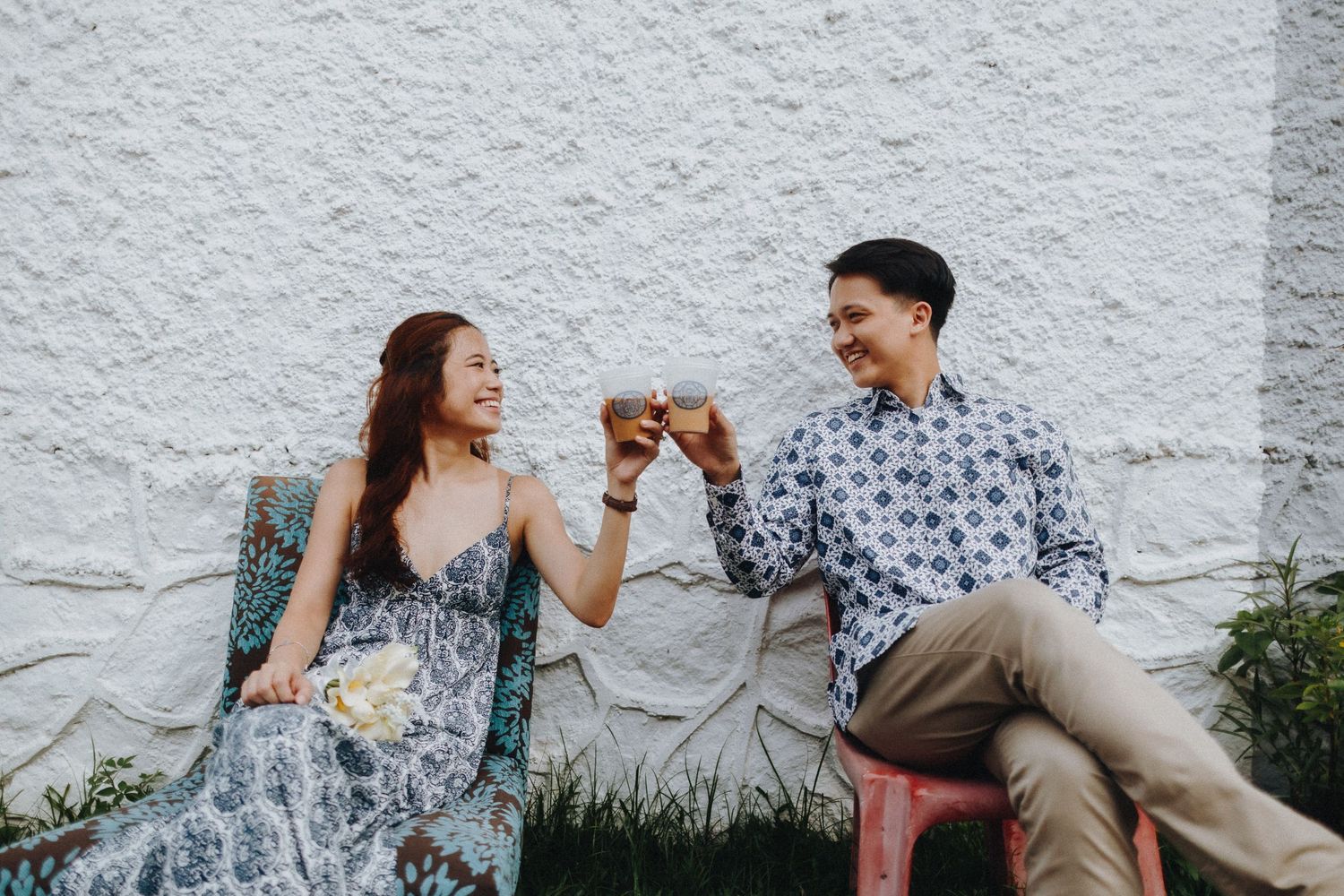 Candid engagement session with couple sitting and laughing together against white wall.
