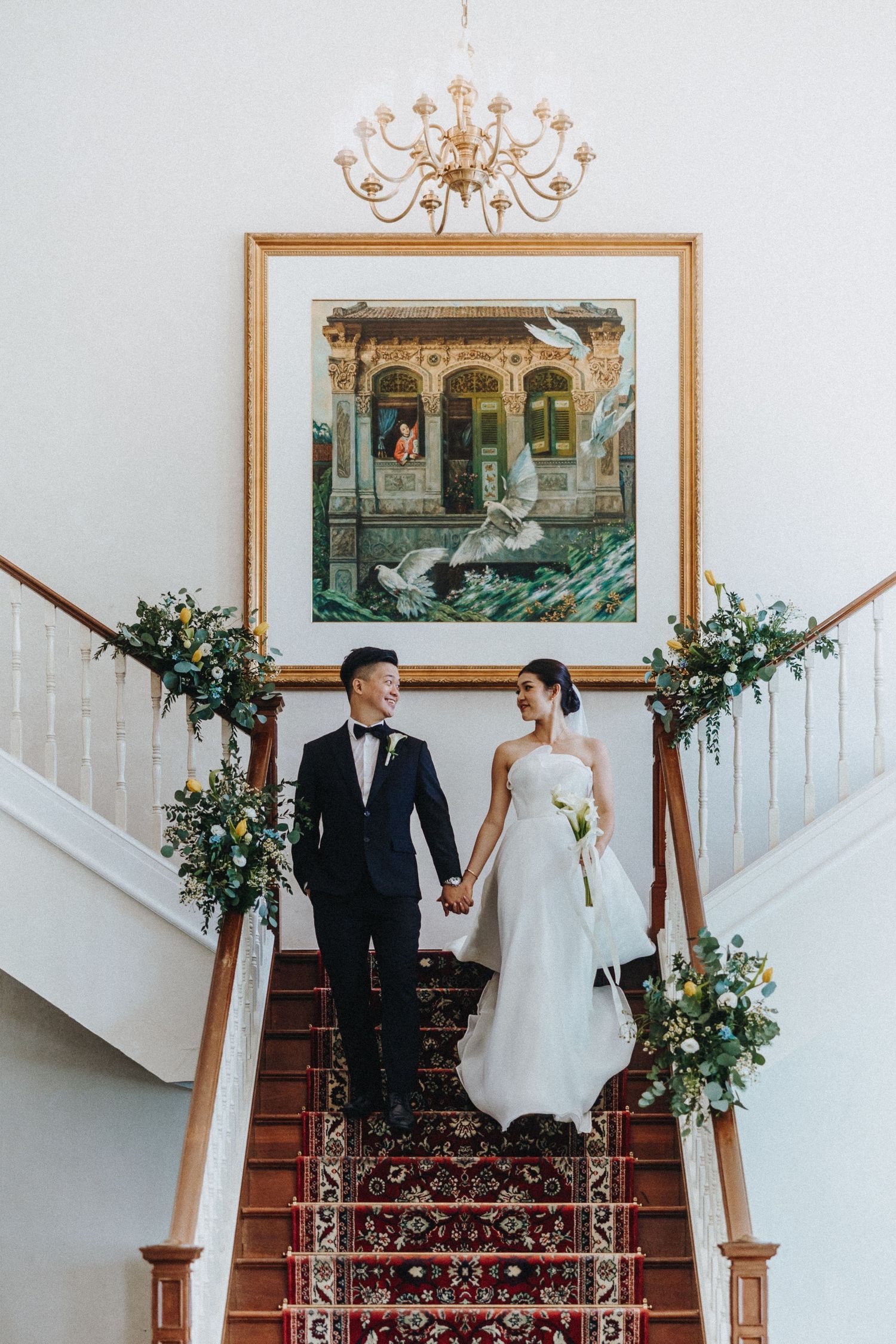 Newlyweds descend ornate staircase decorated with greenery beneath framed artwork and chandelier.