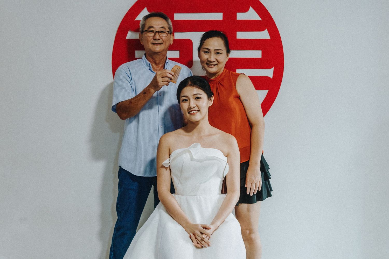 Three people pose together in front of a red Chinese double happiness symbol during a wedding photoshoot.