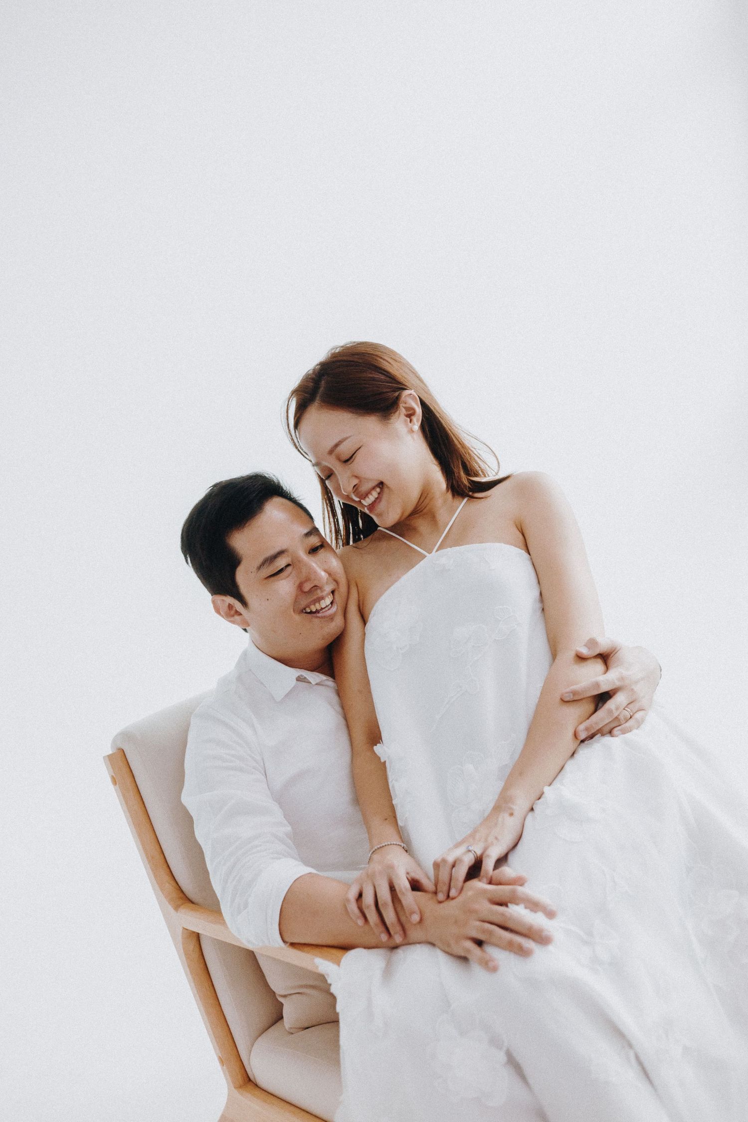 A tender embracing couple in white wedding attire sitting together against a light background.