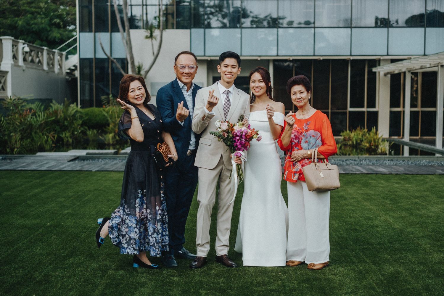 Wedding party poses on a manicured lawn in front of a modern glass building during an outdoor celebration.