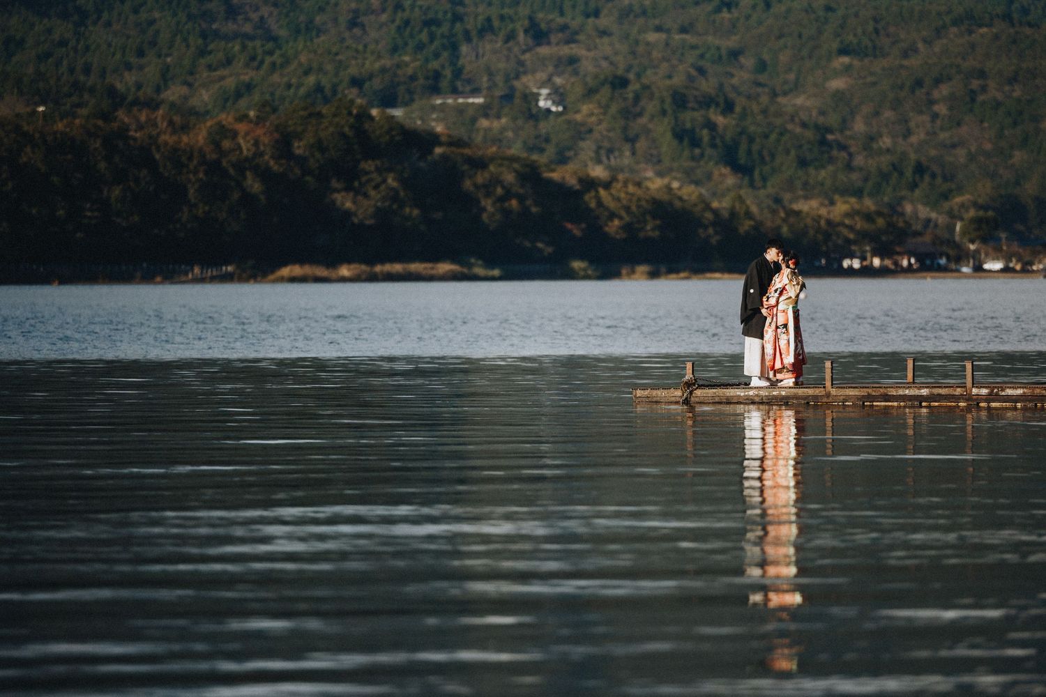 Couple stands on wooden dock surrounded by mountains and clear lake waters at sunset.