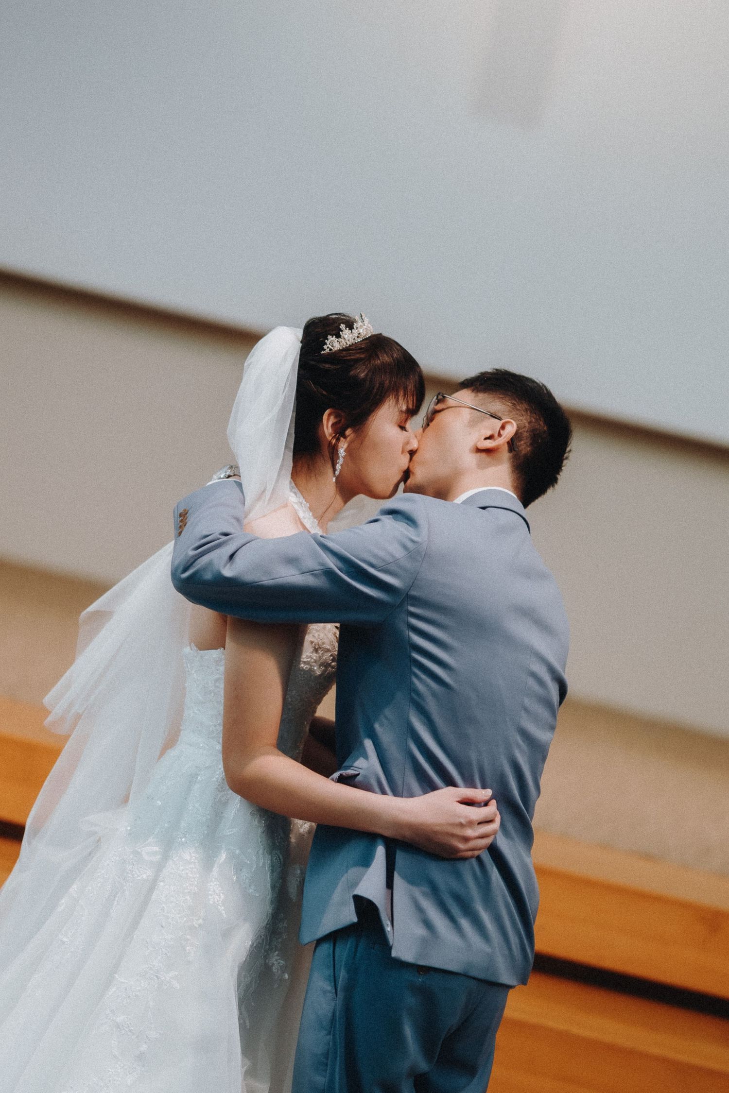 A wedding couple shares a romantic kiss in front of a simple background while wearing traditional wedding attire.
